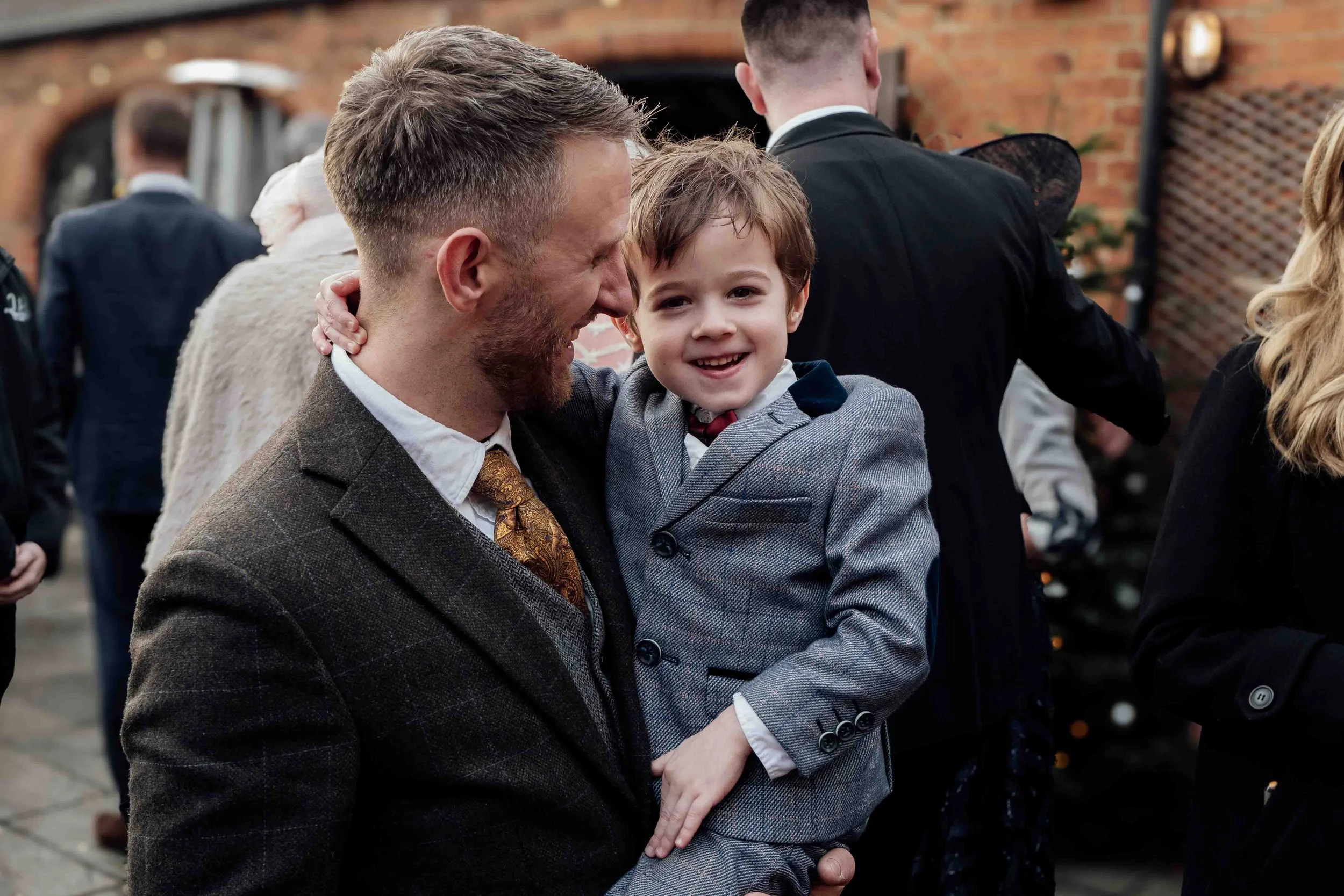 A man holding a young boy at an outdoor social gathering. The man is smiling and looking at the boy, who is smiling directly at the camera. Both are dressed in formal attire, with the man in a brown suit and tie and the boy in a gray suit, tie, and w