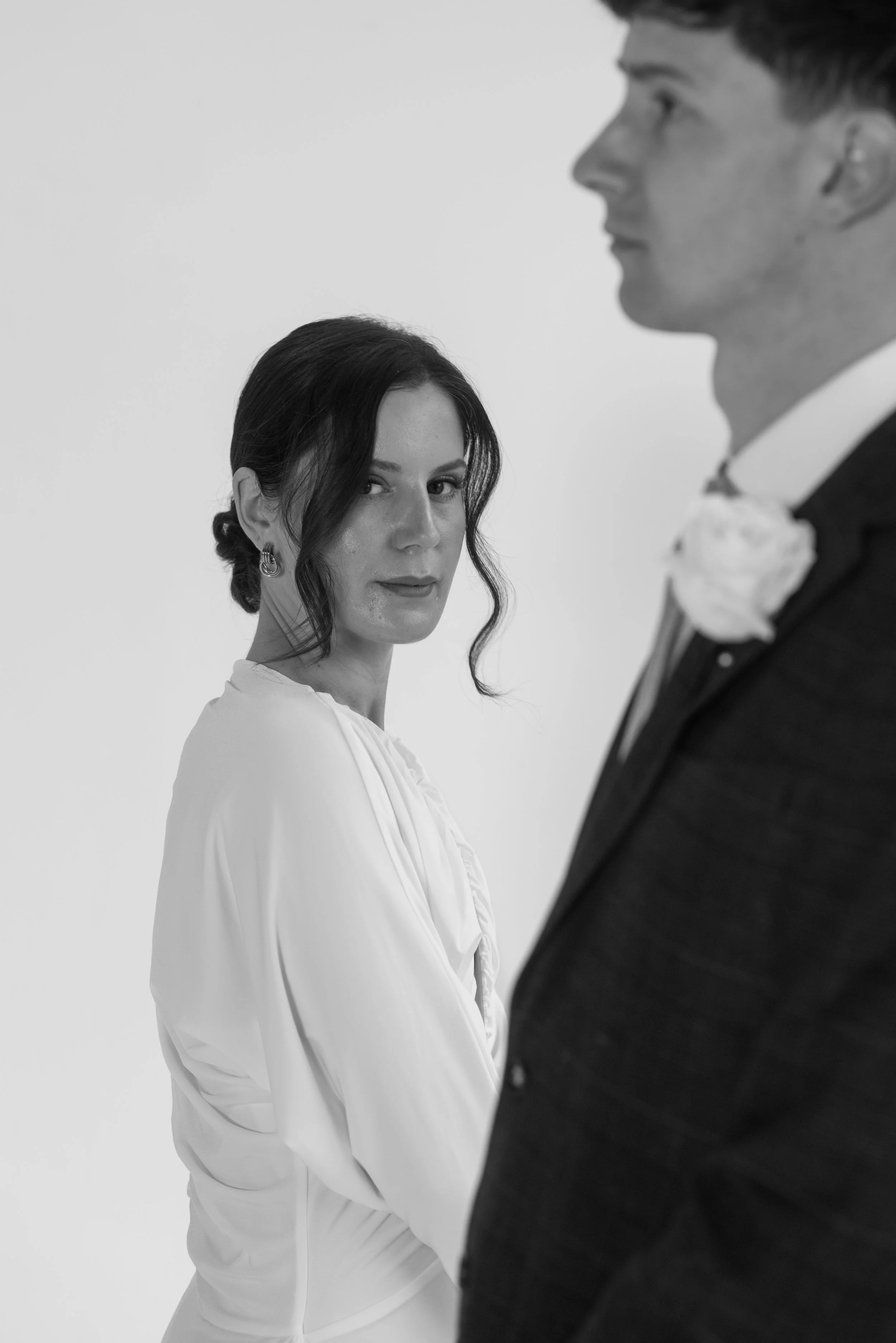 A black-and-white photo of a woman in a white dress looking at a man in a tuxedo with a flower boutonniere, with the woman's face in focus and the man's profile partly visible.