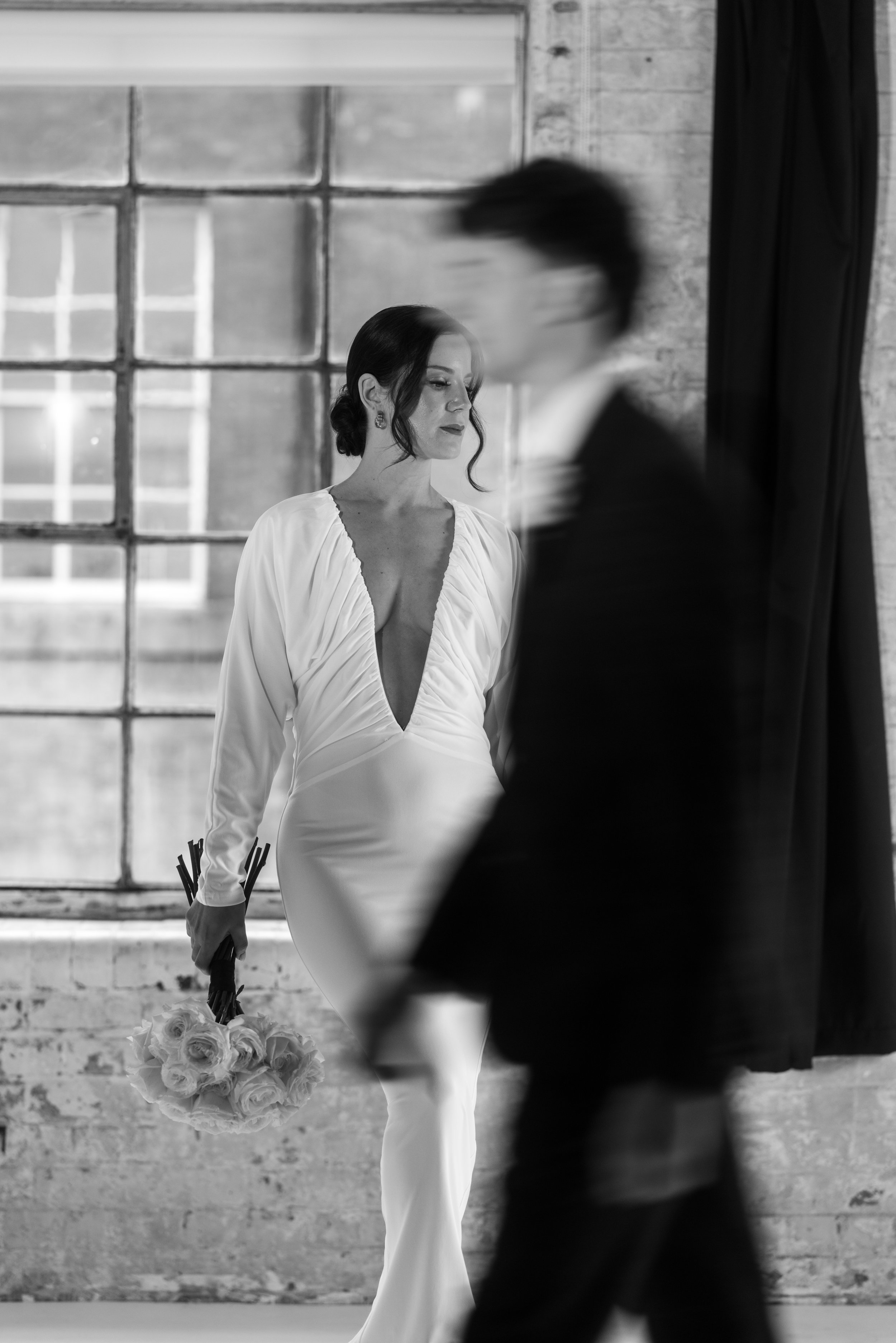 A woman in a white dress holding a bouquet of roses stands near a man in a suit, both inside a room with large industrial-style windows and exposed brick walls, black and white photograph.