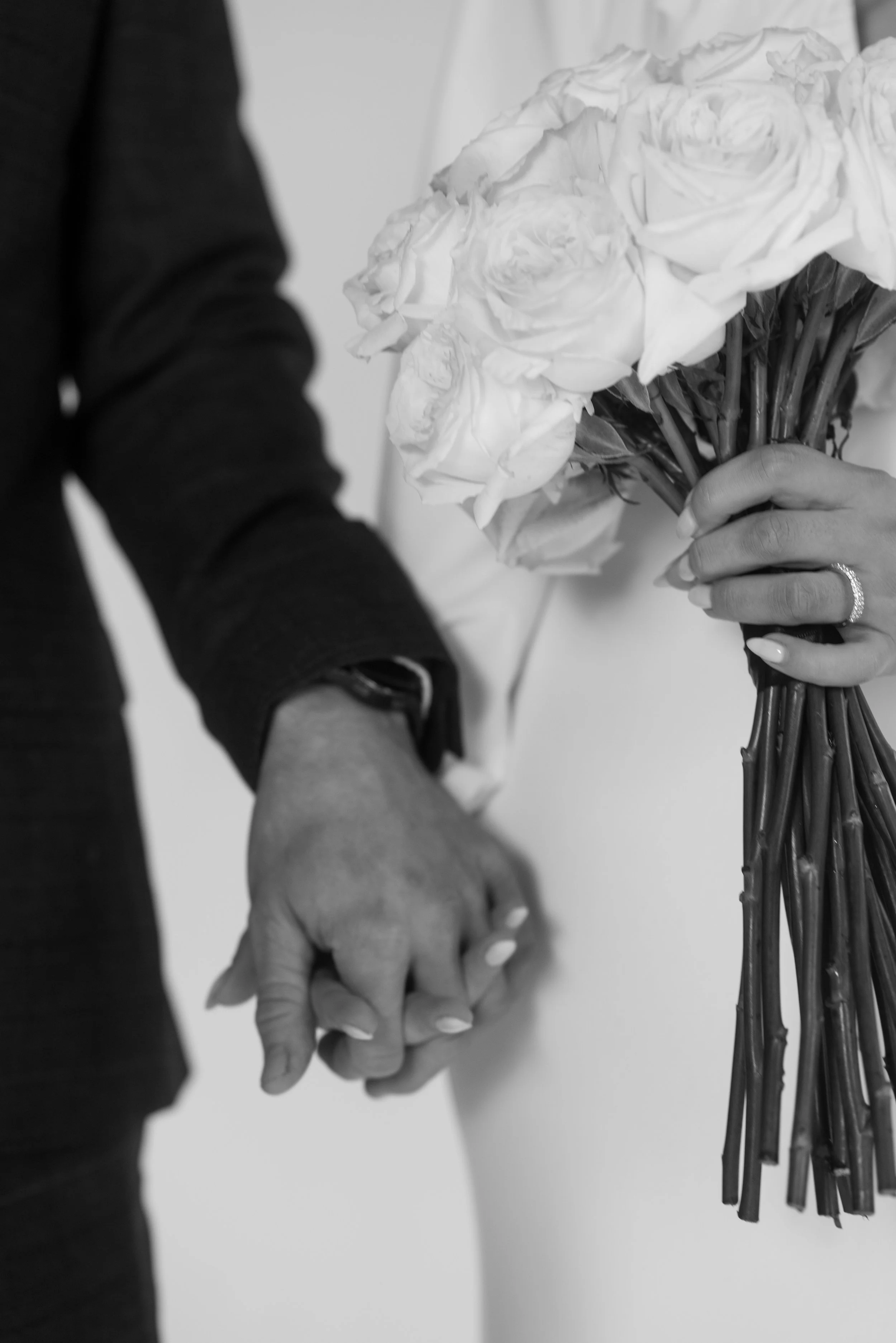 A black-and-white photo of a man and woman holding hands with wedding rings, with the woman holding a bouquet of roses.