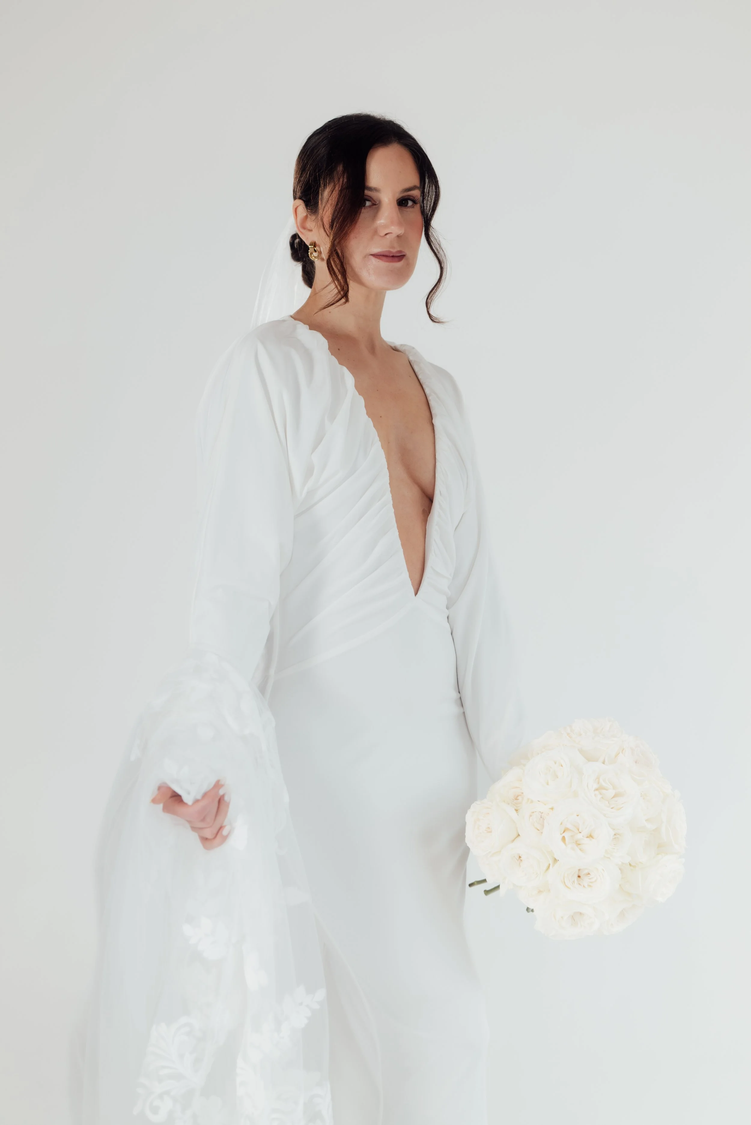 Bride in a white wedding dress holding a bouquet of white roses, standing against a plain white background.