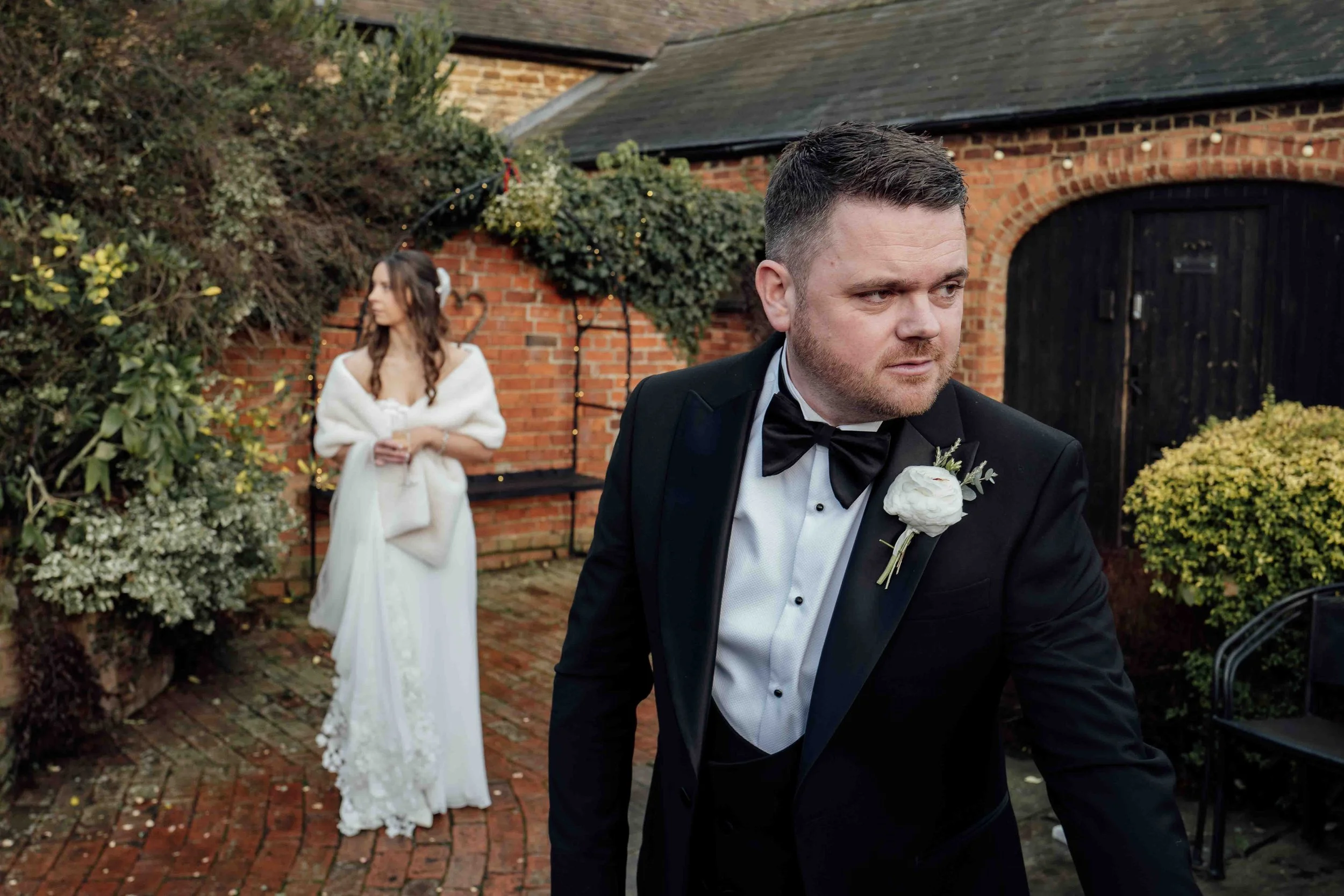 A man in a black tuxedo with a white shirt and black bow tie standing outdoors on a brick patio, looking to his right, with a woman in a white dress and fur stole in the background, holding a glass, in front of a brick wall with greenery and string l
