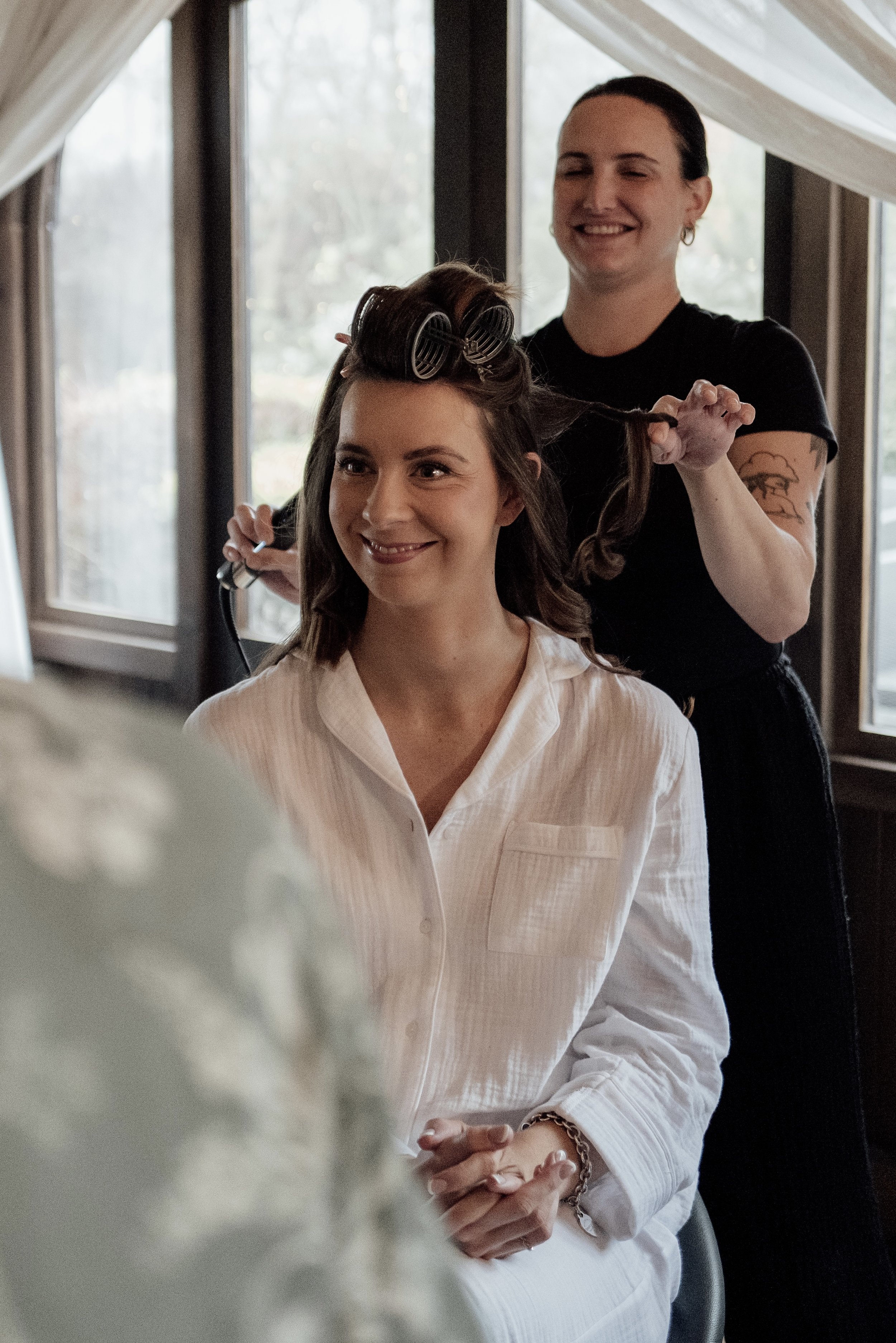 A woman with styled hair sitting in a salon chair, smiling, as a hairstylist with rollers and curlers in her hair styles her hair.