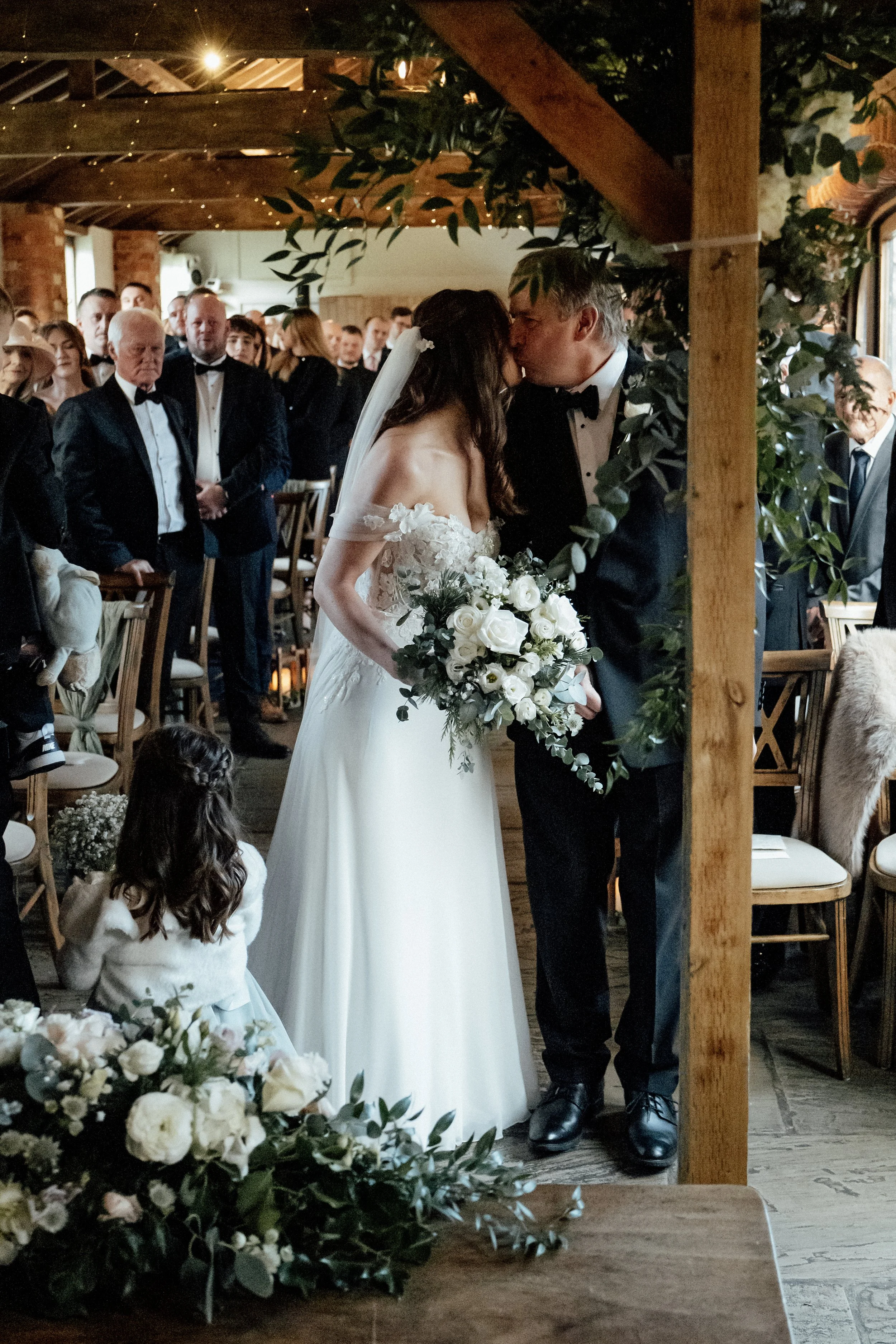A bride and groom kissing during their wedding ceremony indoors, surrounded by seated guests. The bride is holding a bouquet of white roses and greenery, wearing an off-shoulder wedding gown with lace details. The groom is in a black tuxedo with a bo