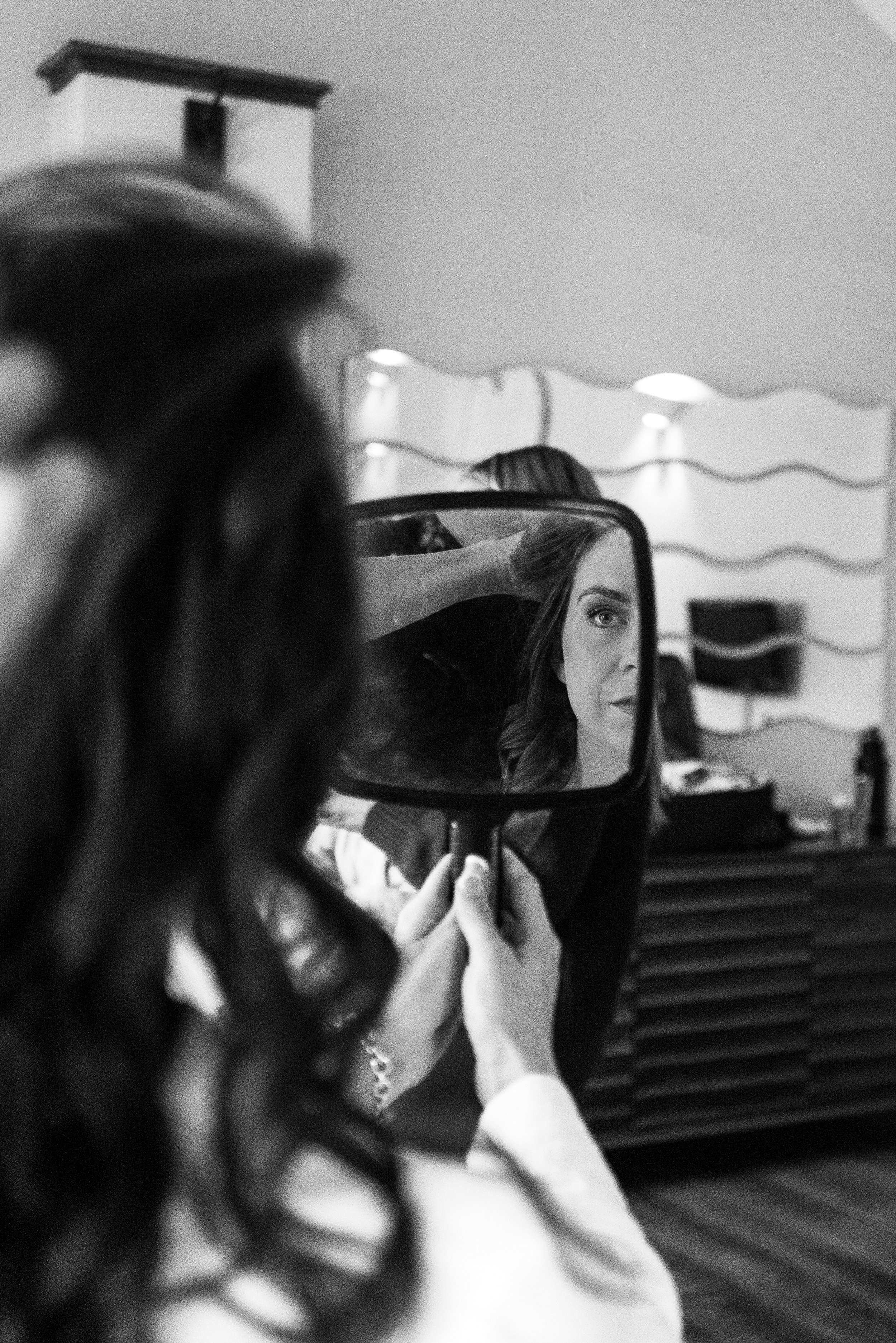 A woman with wavy hair looks into a mirror held by another person, who appears to be helping her get ready.