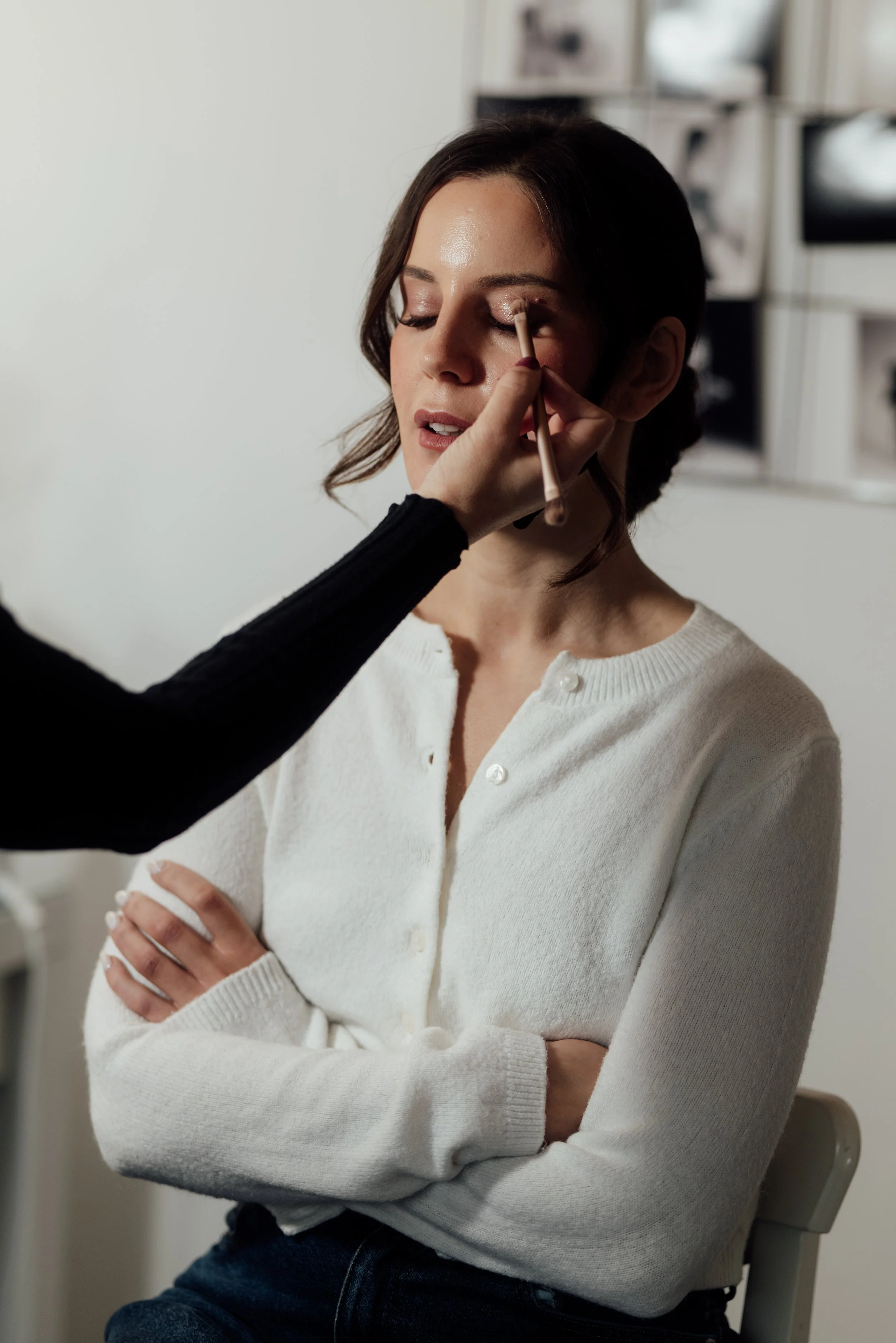 A woman with brown hair and wearing a white sweater gets her makeup done by a makeup artist.