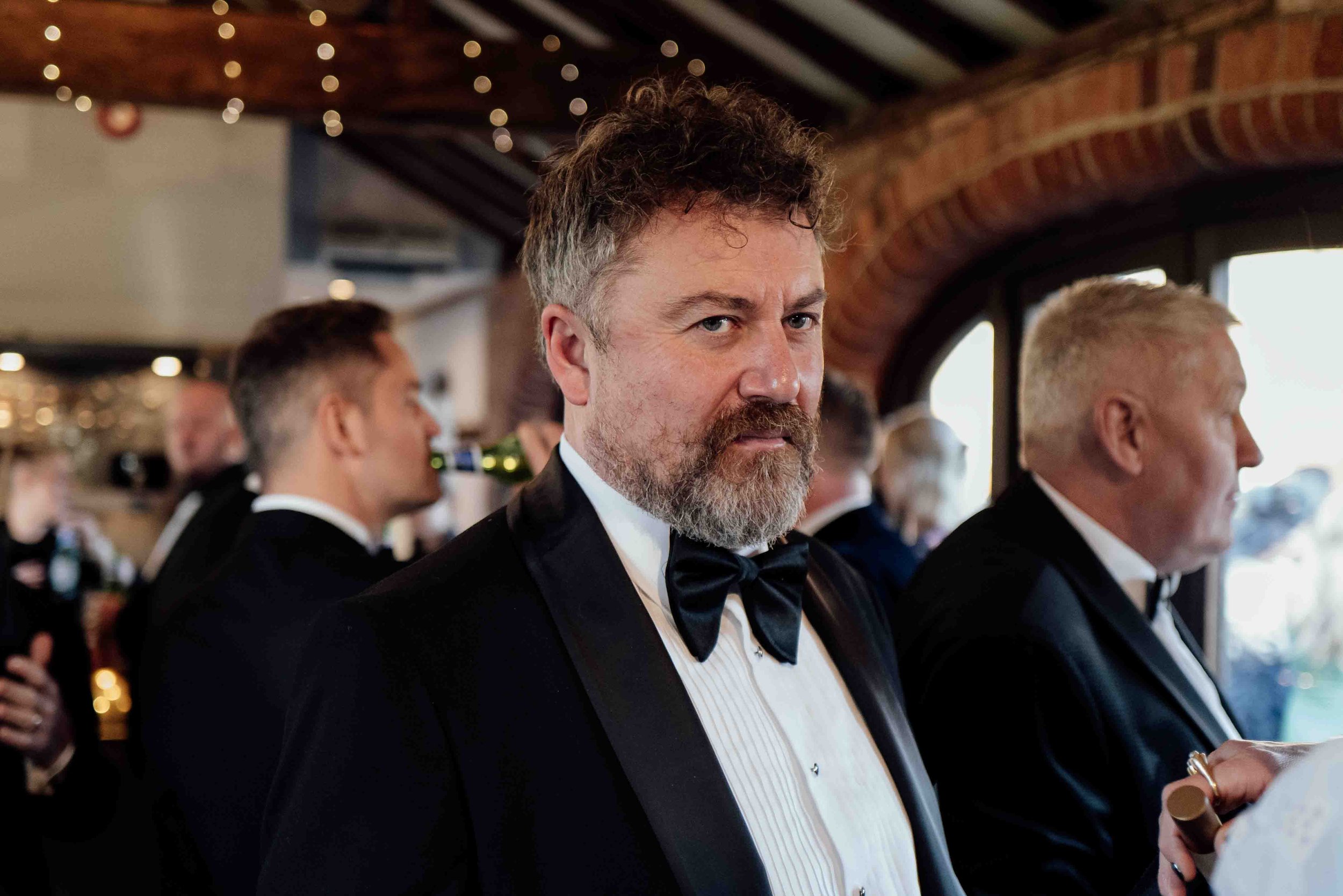 A man with a beard and curly hair, wearing a tuxedo with a bow tie, at a formal event or restaurant, surrounded by other people also dressed in formal attire, in a warmly lit setting with brick walls and large windows.