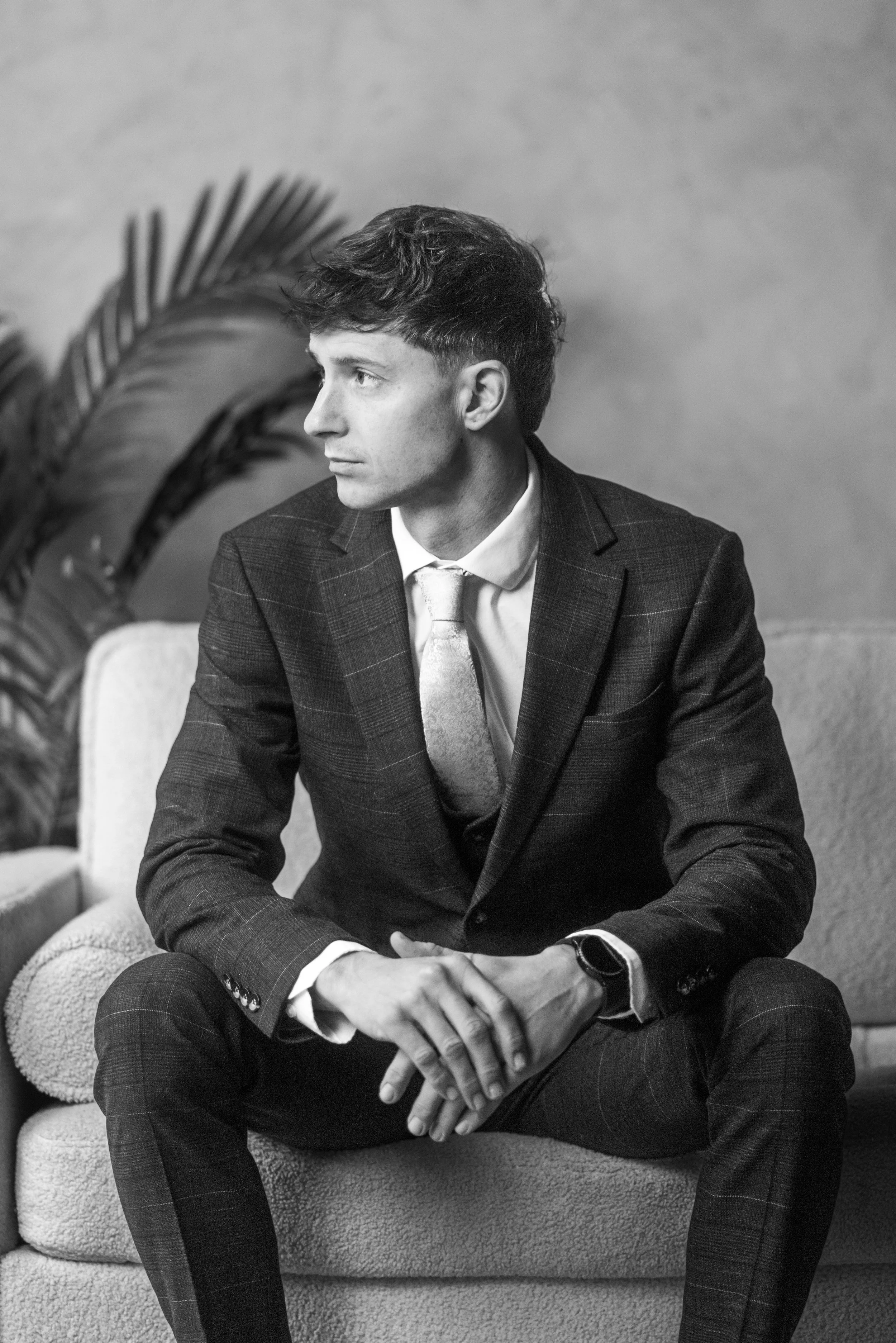 A young man in a dark checkered suit, light shirt, and patterned tie, sitting on a sofa with his hands clasped, looking to the side. There are houseplants in the background.