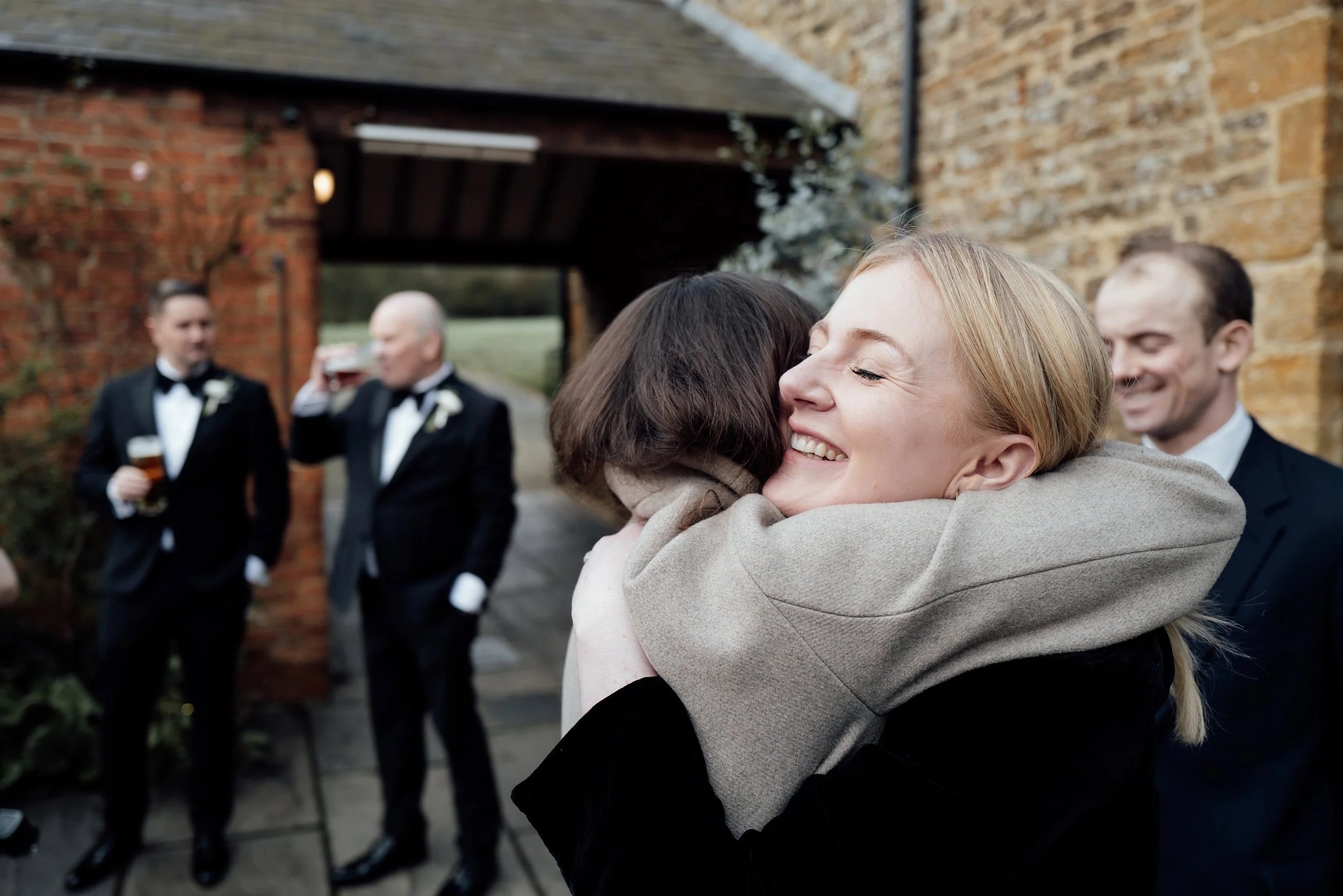 People hugging at a wedding reception outdoors, with two men in tuxedos holding drinks in the background.