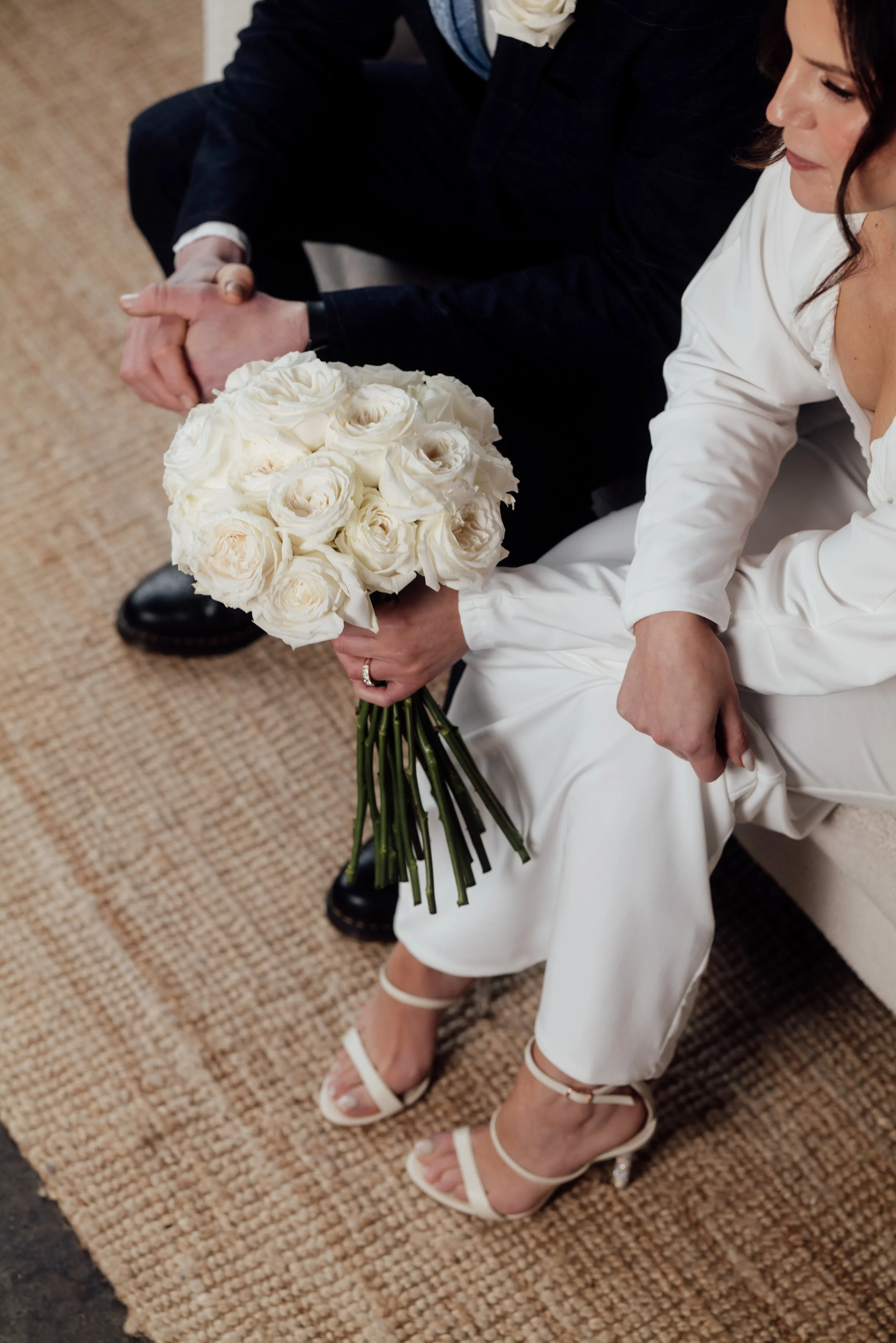 A bride in white wedding dress and heels holding a bouquet of white roses, sitting next to a man in dark suit, both clasping hands.