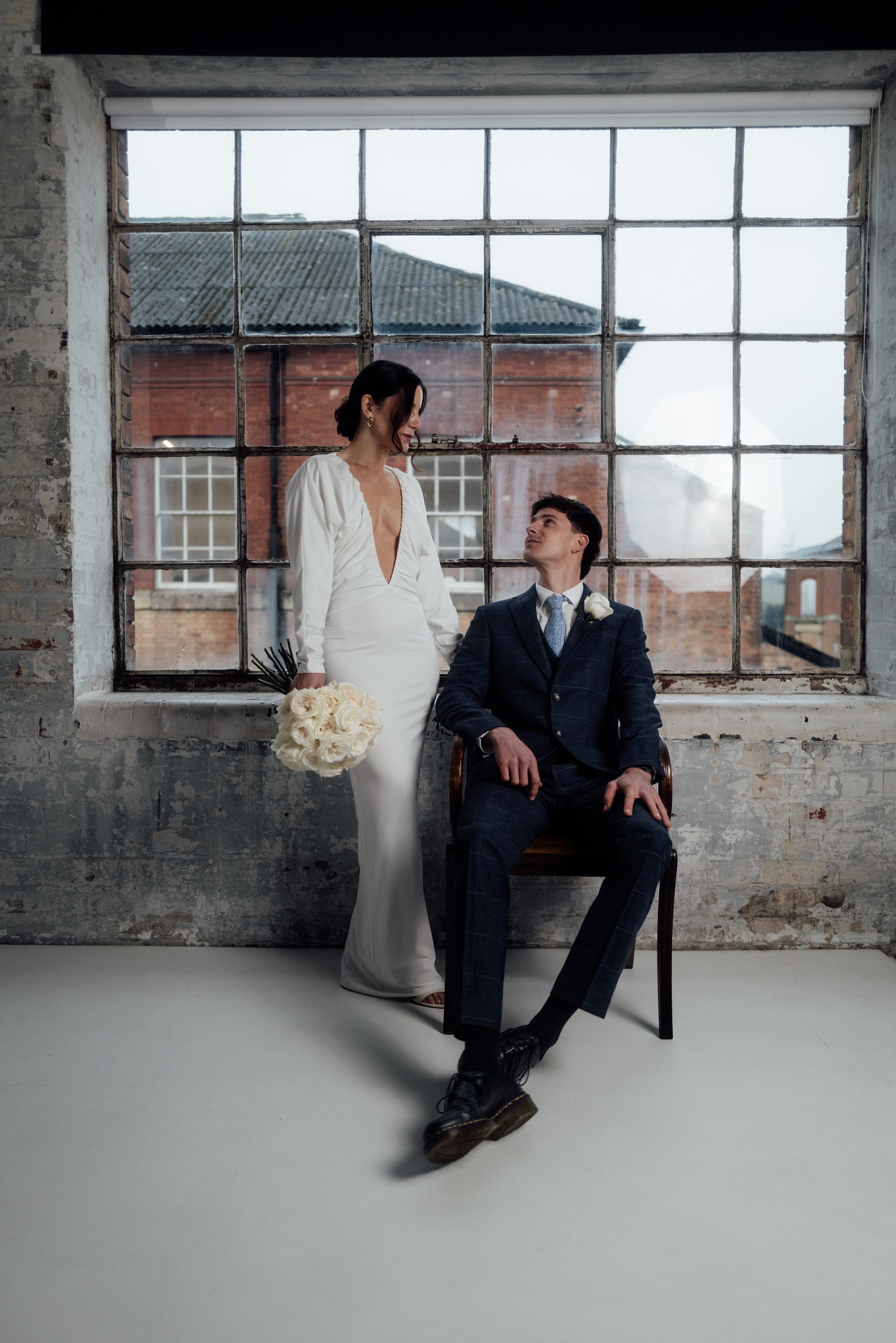 A bride in a white wedding dress holding a bouquet of white roses stands next to a seated groom in a plaid suit, inside an industrial-style room with a large window and exposed brick walls.