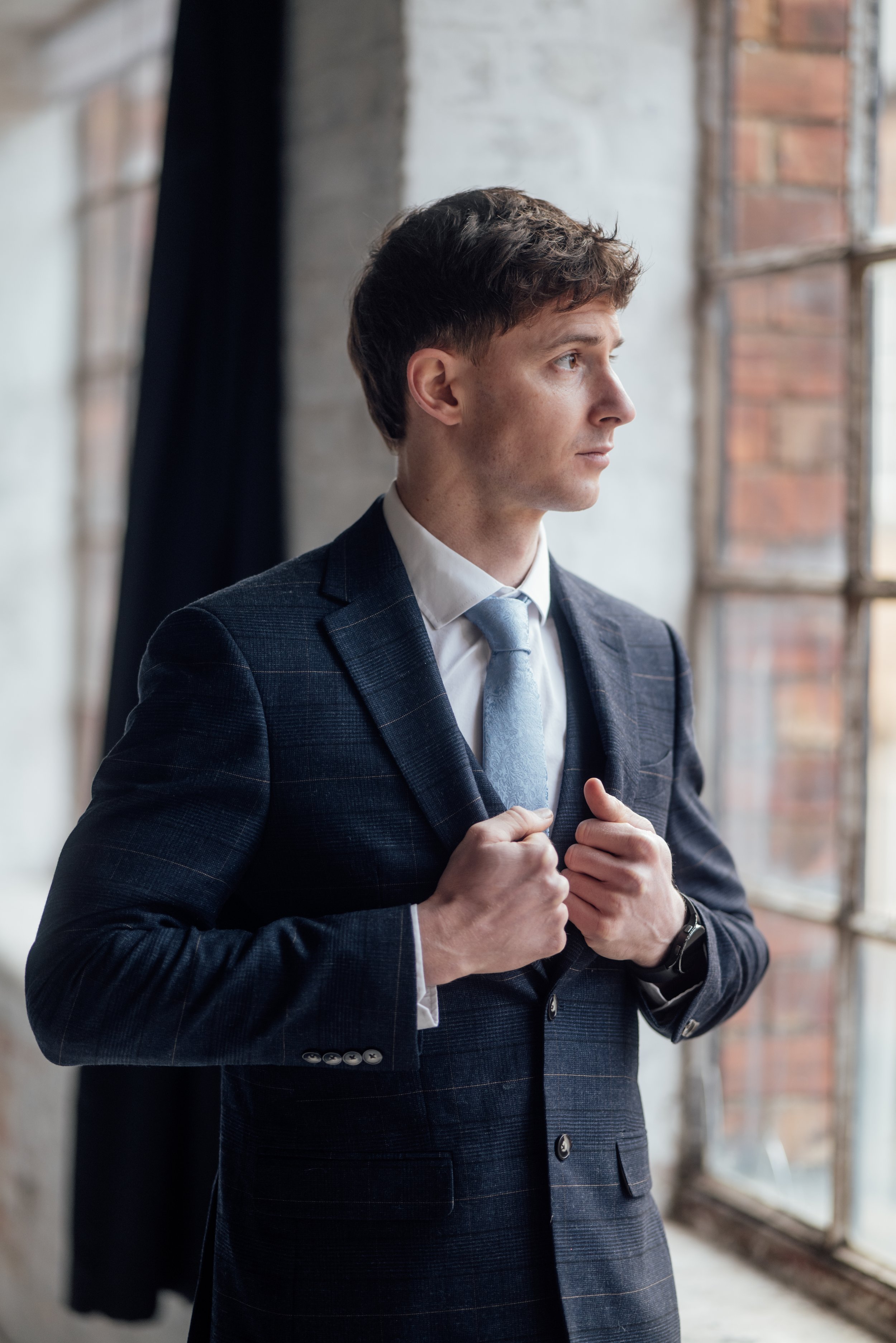 A young man in a dark, checkered suit with a white shirt and light blue tie looking contemplative, standing near a large window in an industrial-style room.