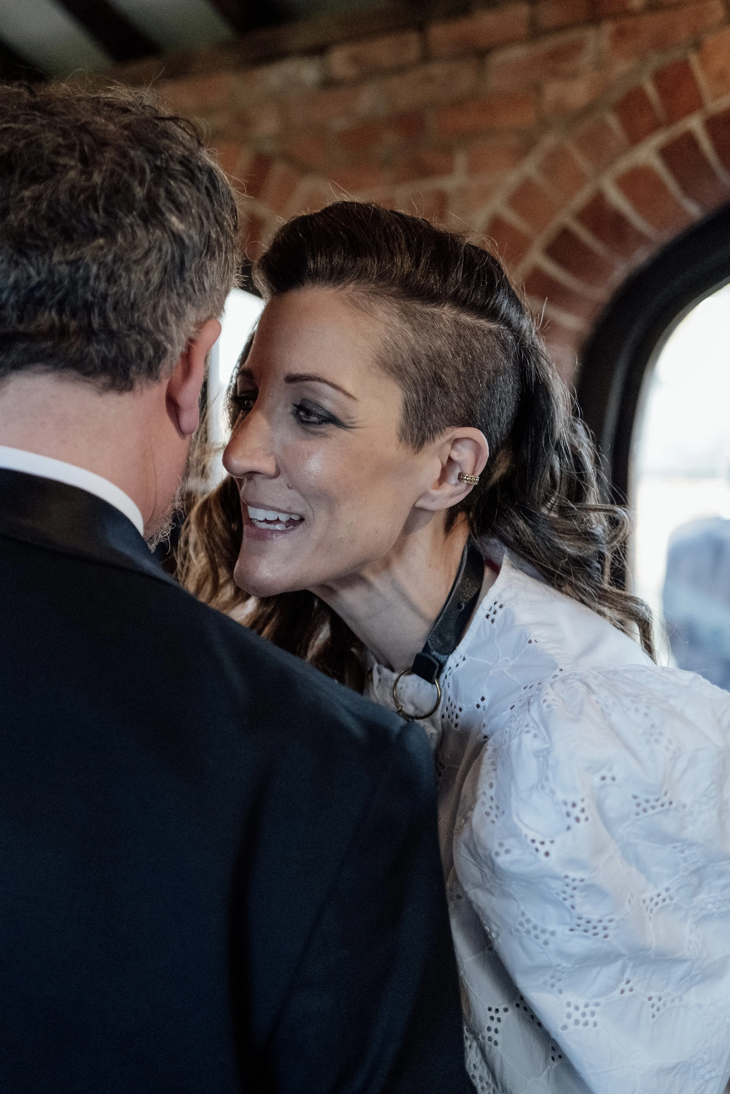 A woman with a short, undercut hairstyle and a white embroidered blouse is smiling and leaning in towards a man in a black tuxedo, inside a brick building near a window.