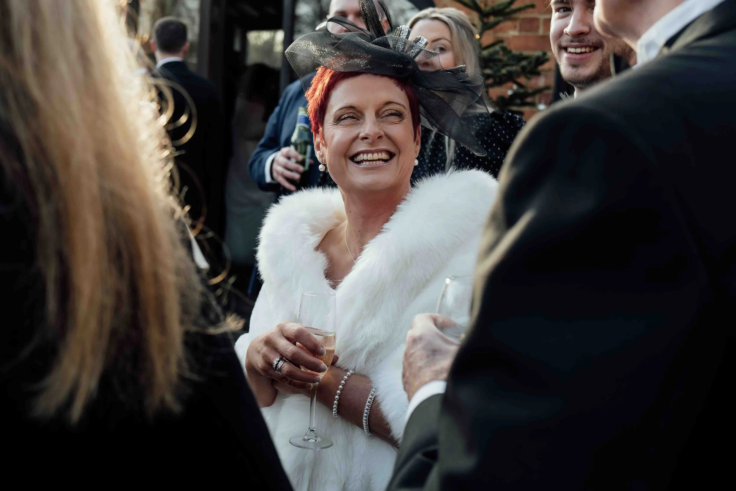 A woman with short red hair, wearing a white fur coat and a black fascinator, smiling and holding a glass of champagne at a social gathering.