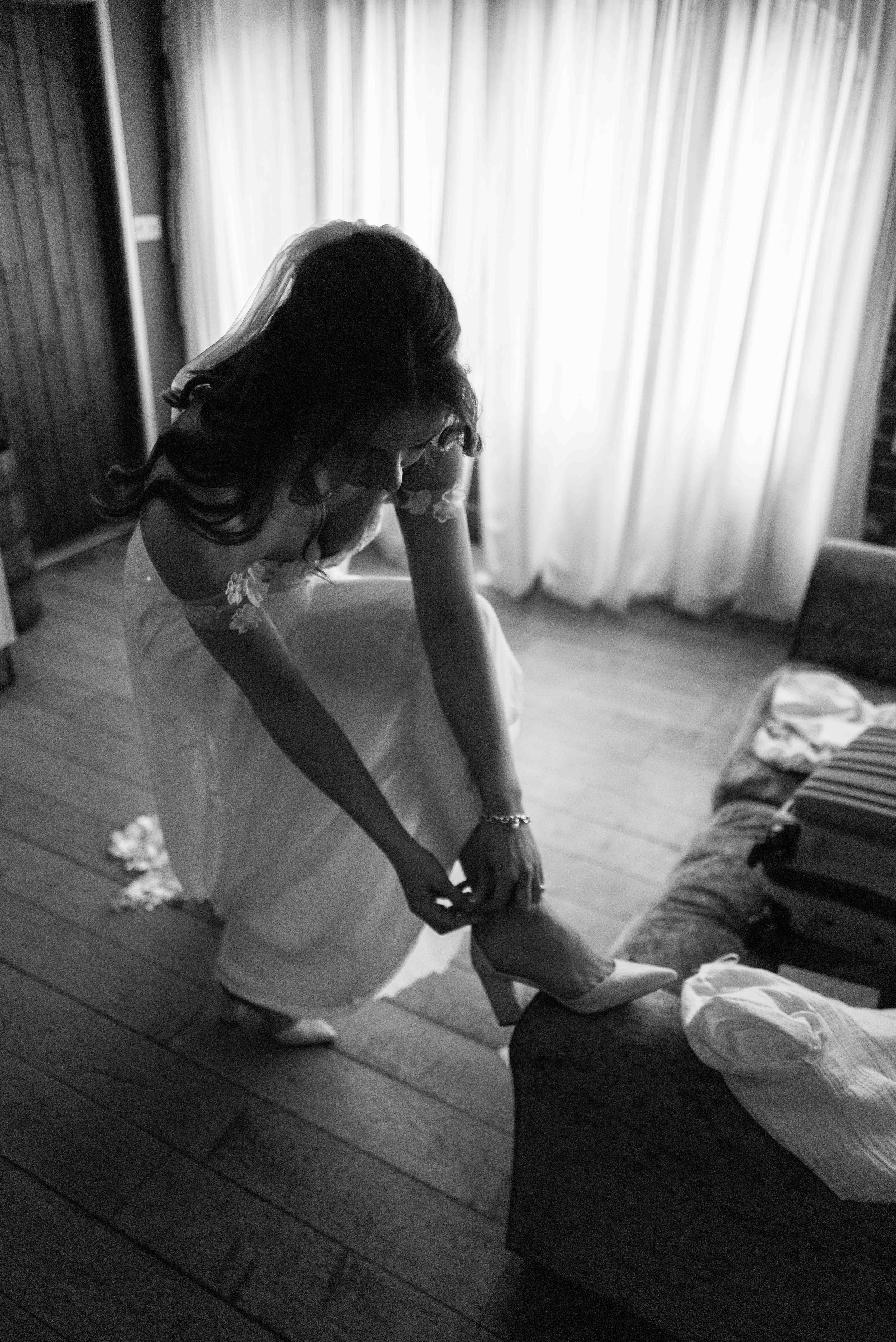 A woman in a wedding dress puts on high-heeled shoes in a room with curtains and a sofa.