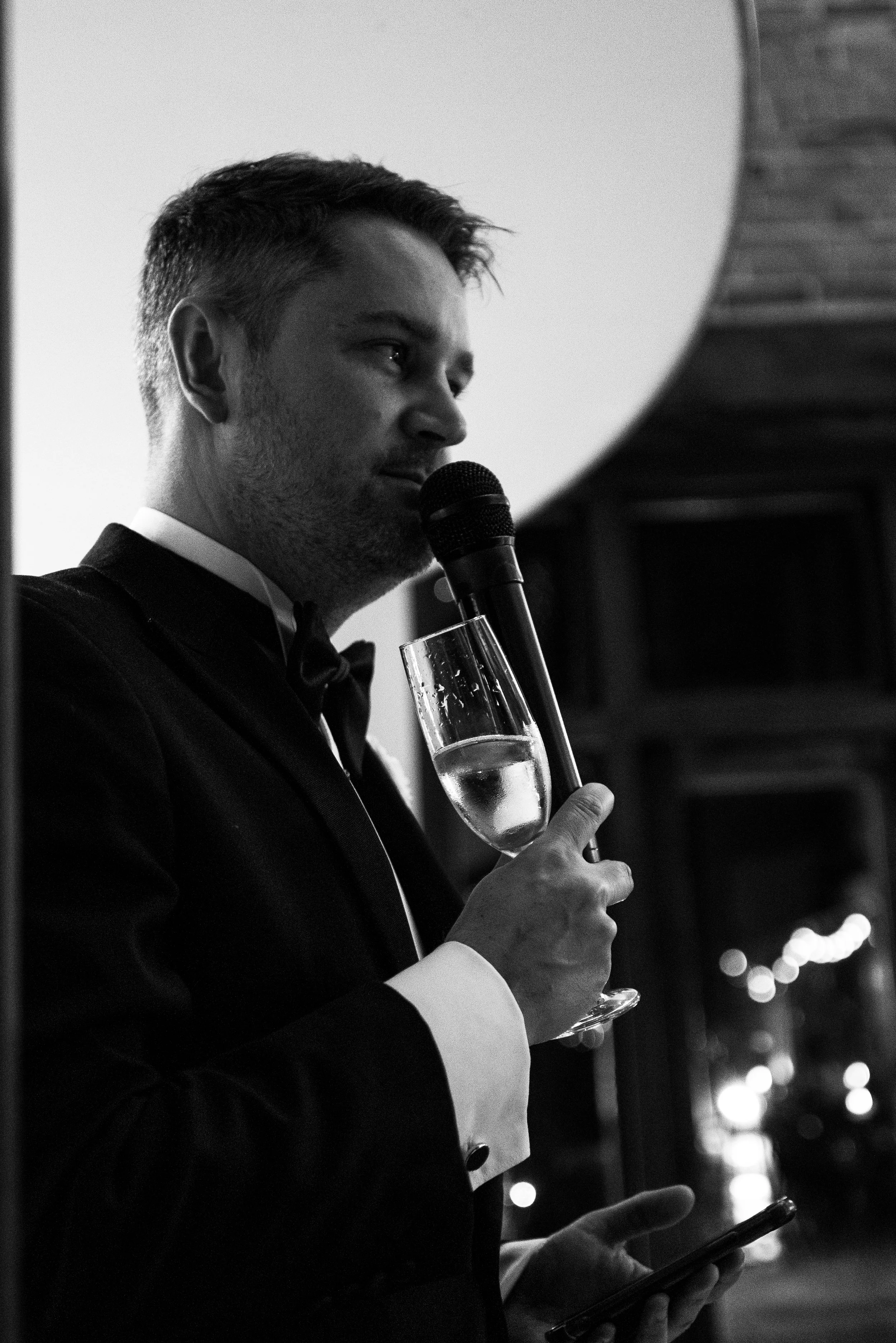 A man in a tuxedo holding a glass of champagne and a microphone, standing indoors at a formal event.
