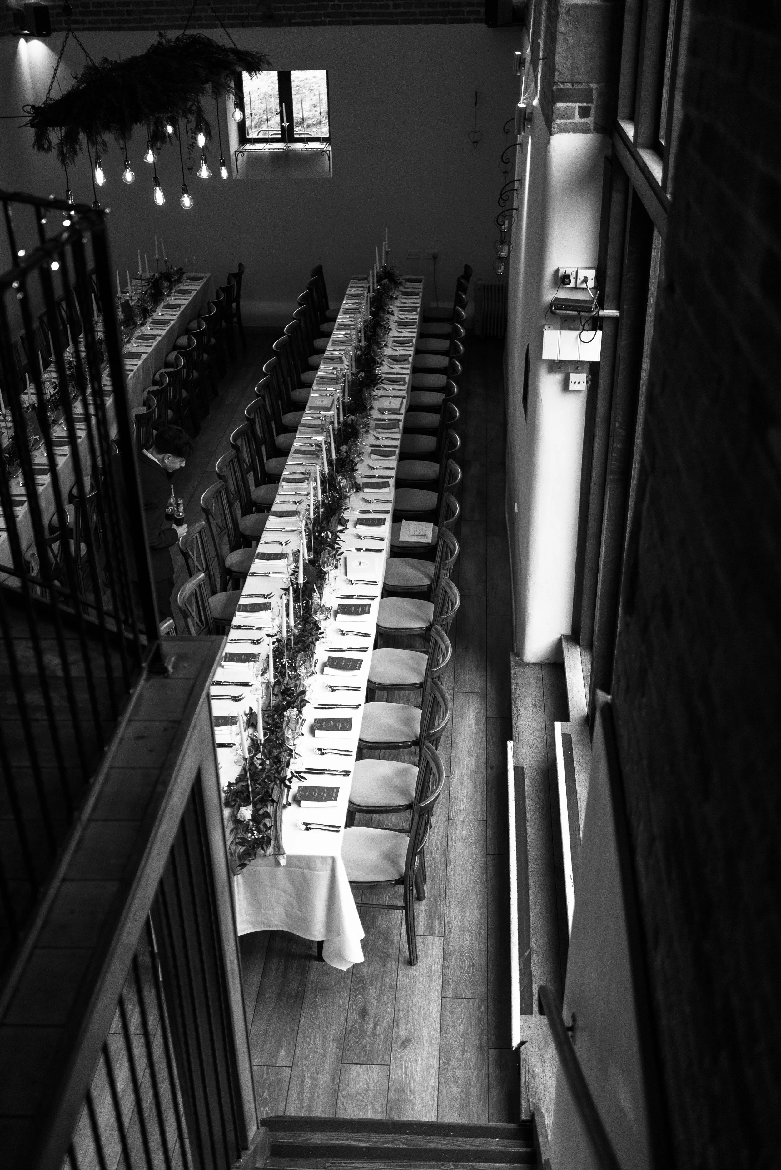 Long banquet table set up for a formal event with place settings, glassware, and floral centerpieces, viewed from above in a rustic-style dining area with wooden floors and hanging light fixtures.