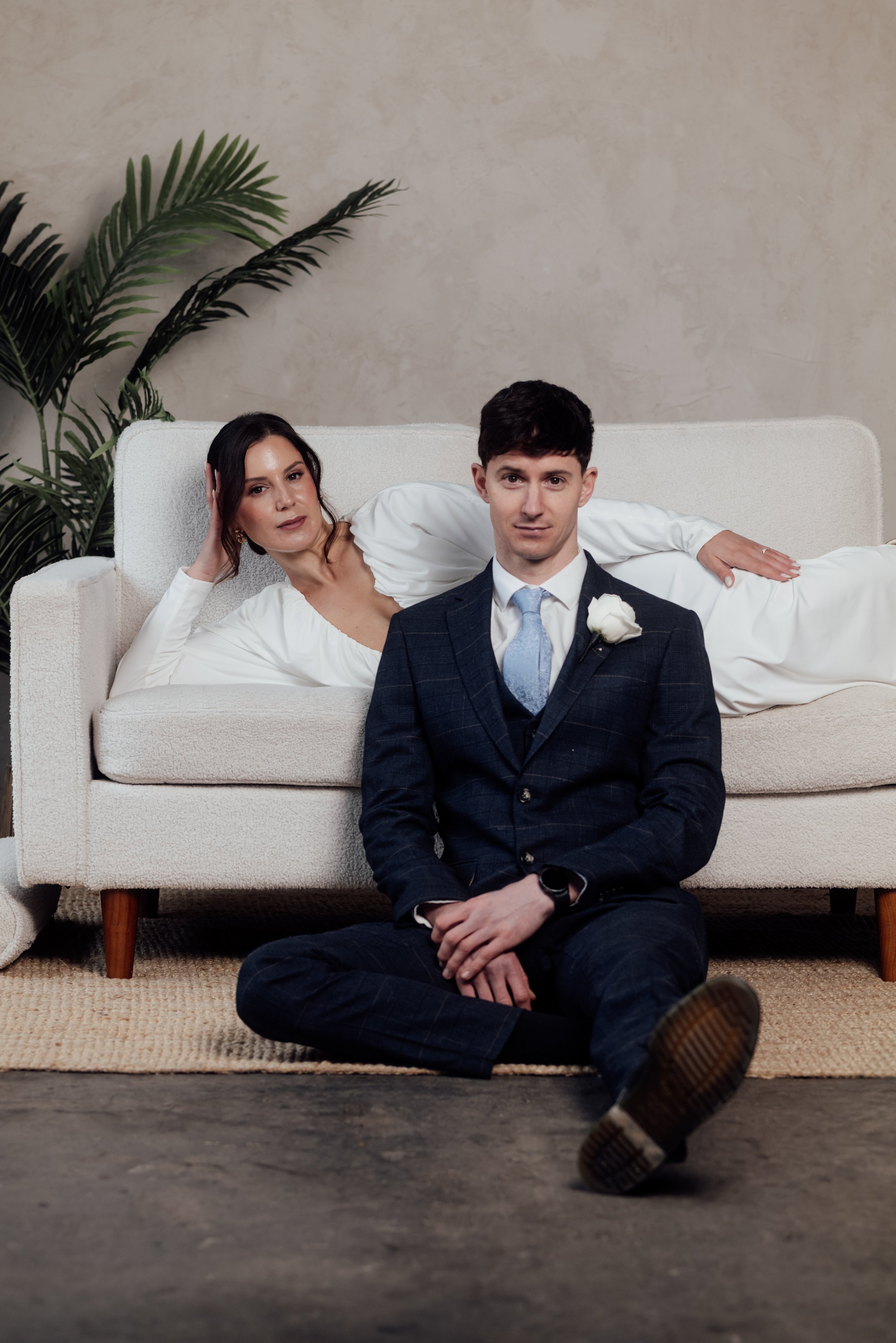 A man in a suit sitting on the floor in front of a woman lying on a white couch, both indoors with a plant in the background.