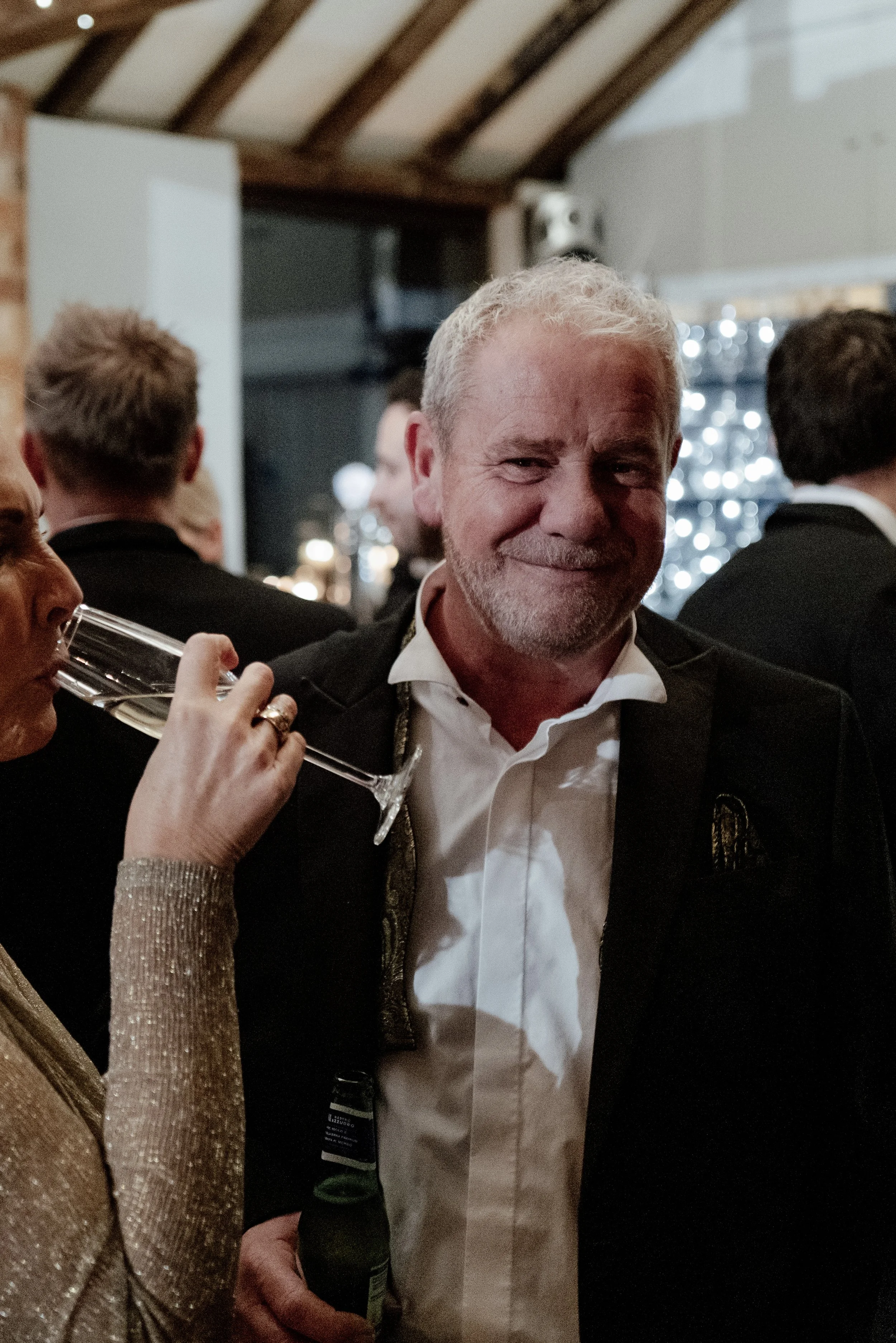 A smiling man in a tuxedo at a social event, holding a beer bottle, with a woman next to him drinking champagne, in a festive indoor setting with other people in the background.
