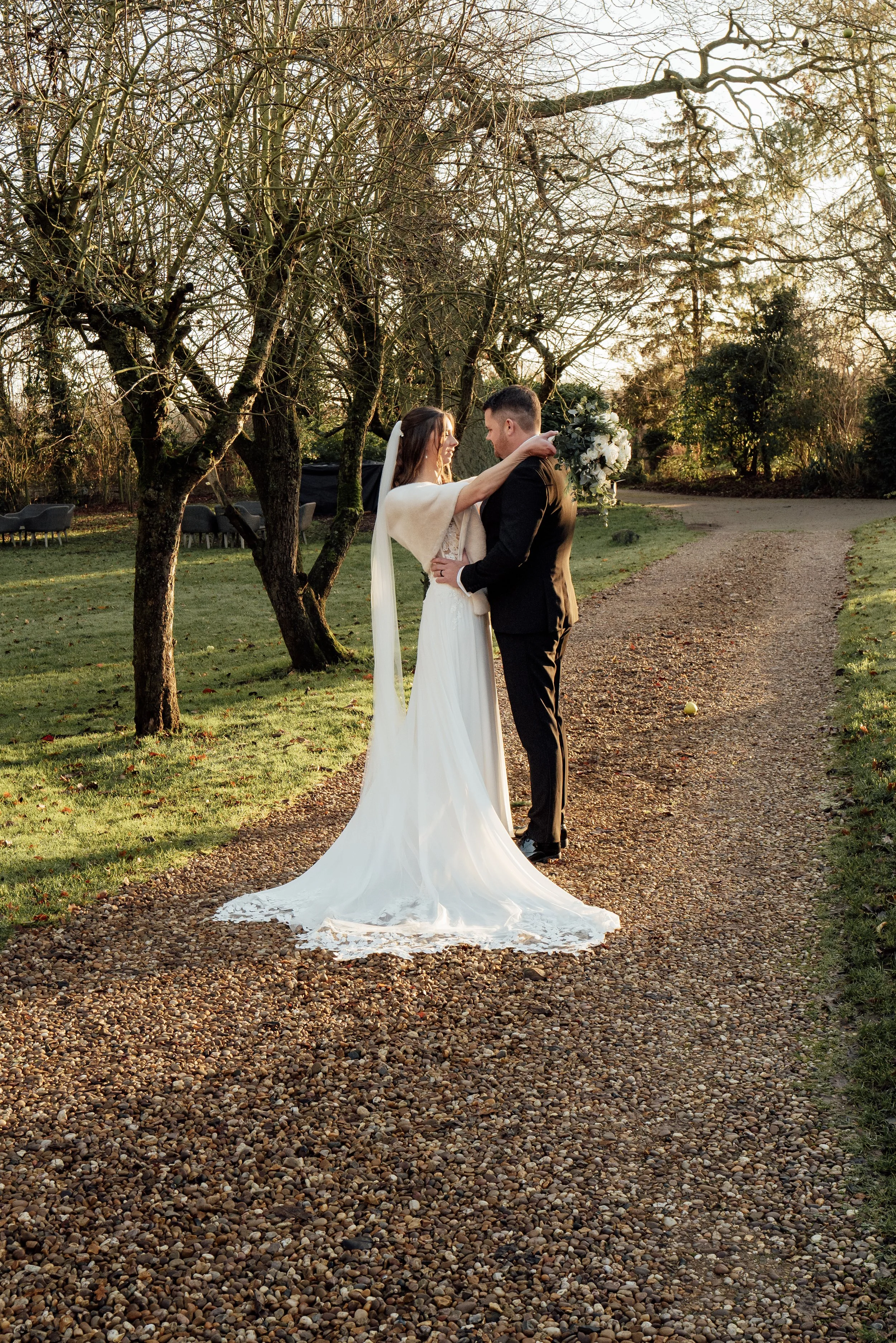 A bride and groom stand close together on a gravel path in a garden, embracing during sunset. The bride wears a white wedding dress with a long train, and the groom wears a black suit. They are surrounded by leafless trees and green grass.