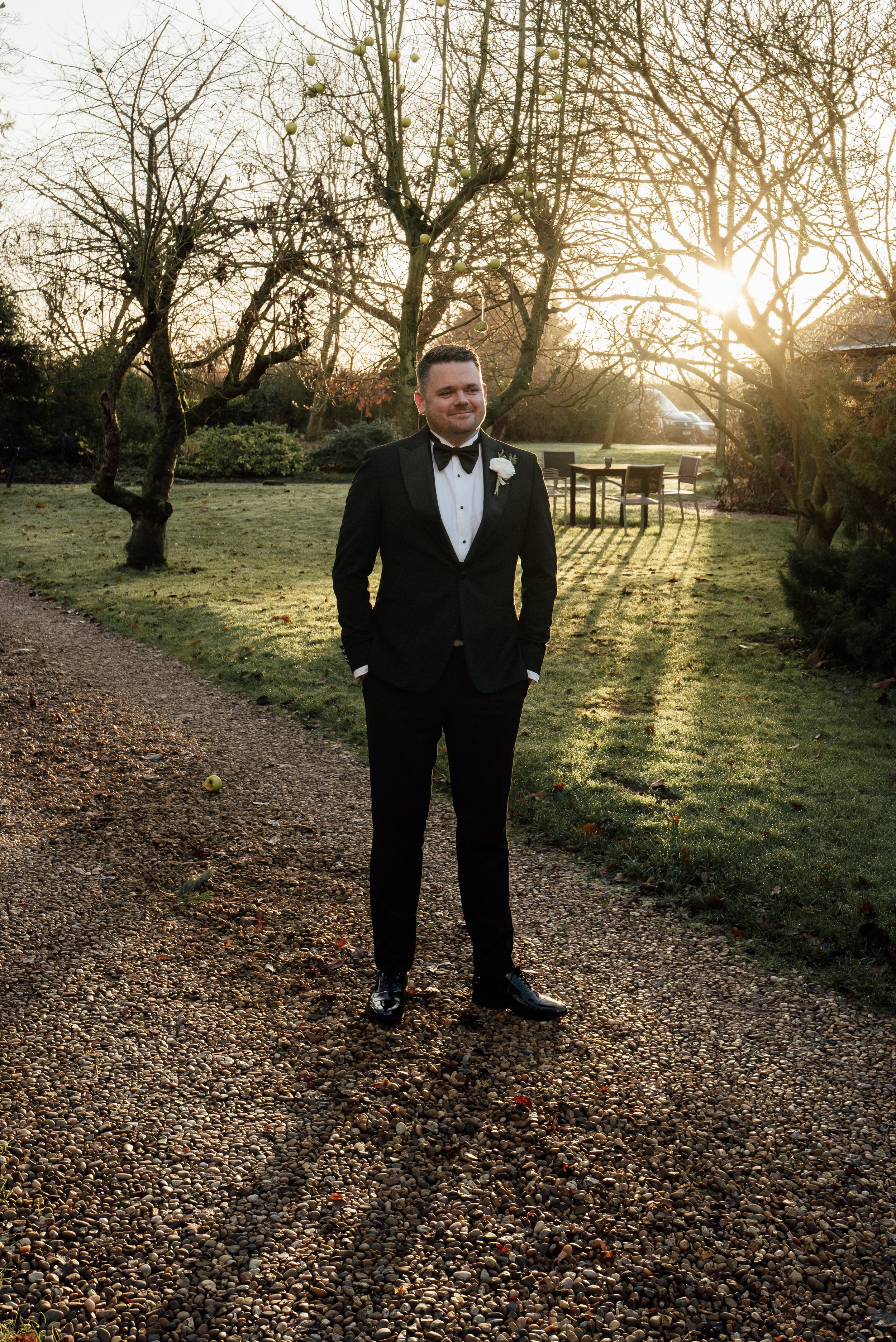 A groom in a black tuxedo with a bow tie and boutonnière stands outdoors on a gravel path with his hands in his pockets. The sun is setting behind leafless trees, casting long shadows and a warm glow. There are outdoor tables and chairs in the backgr
