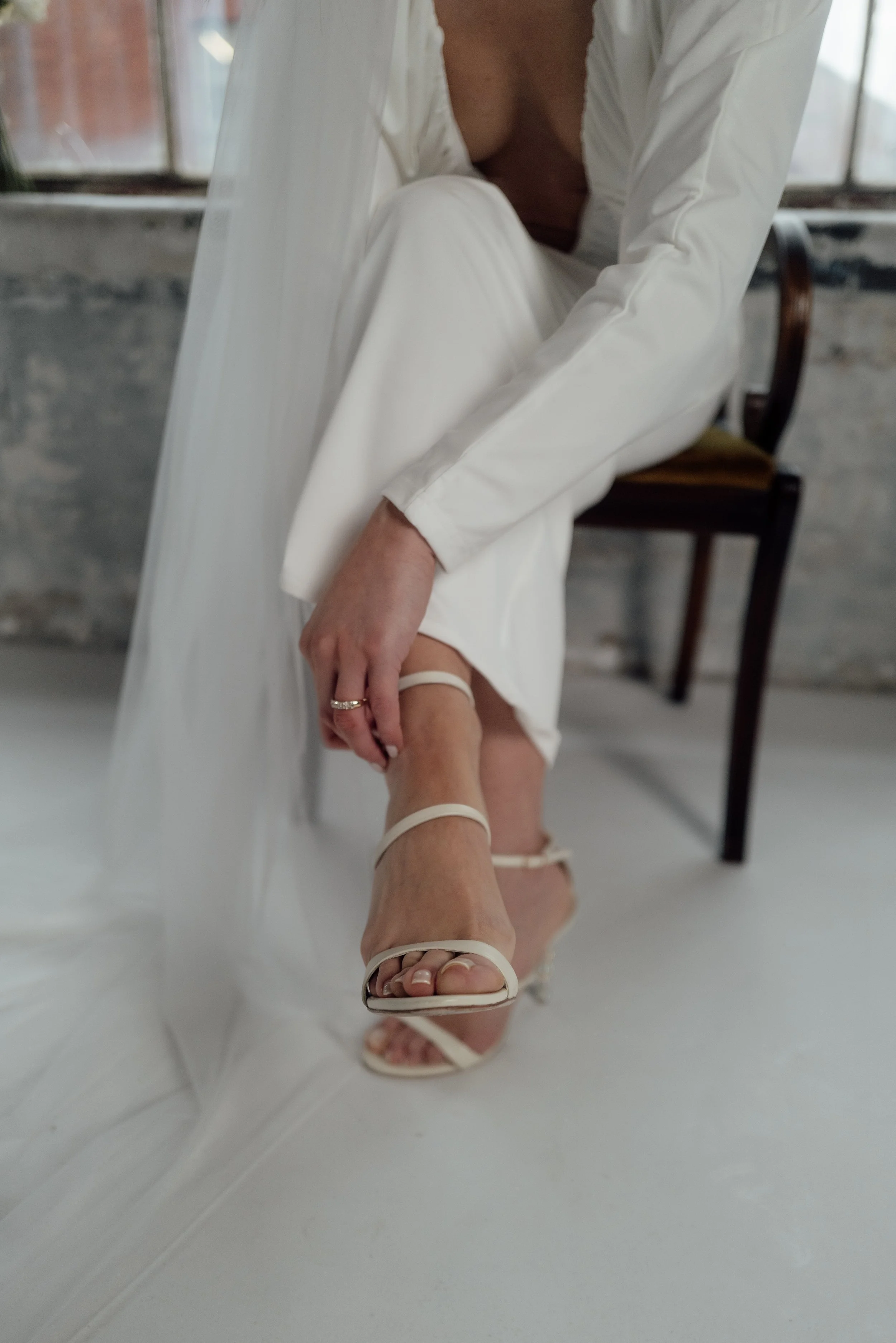 A woman in white attire adjusting her beige high-heeled sandals while sitting on a chair in a minimalist, industrial-style room.