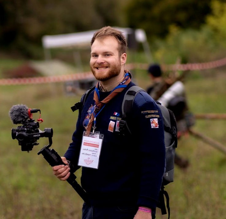 Jeune homme souriant avec un sac à dos, portant une écharpe et un badge, tenant un stabilisateur avec une caméra à l'extérieur dans un environnement naturel. En arrière-plan, une autre personne et une zone délimitée avec une bande de ruban.
