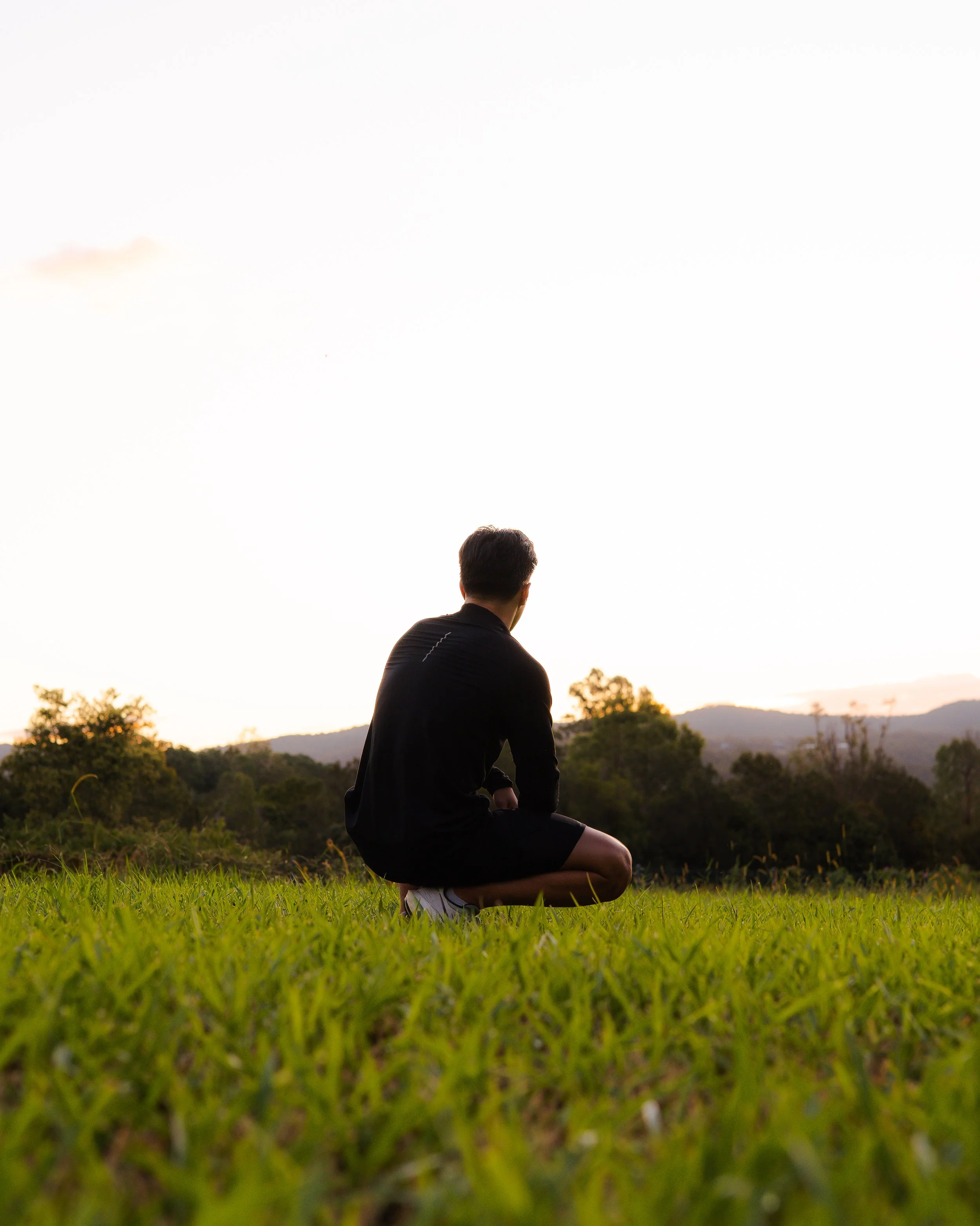 A person in black clothing kneeling on the grass and looking at the sunset in a natural landscape with trees and hills.