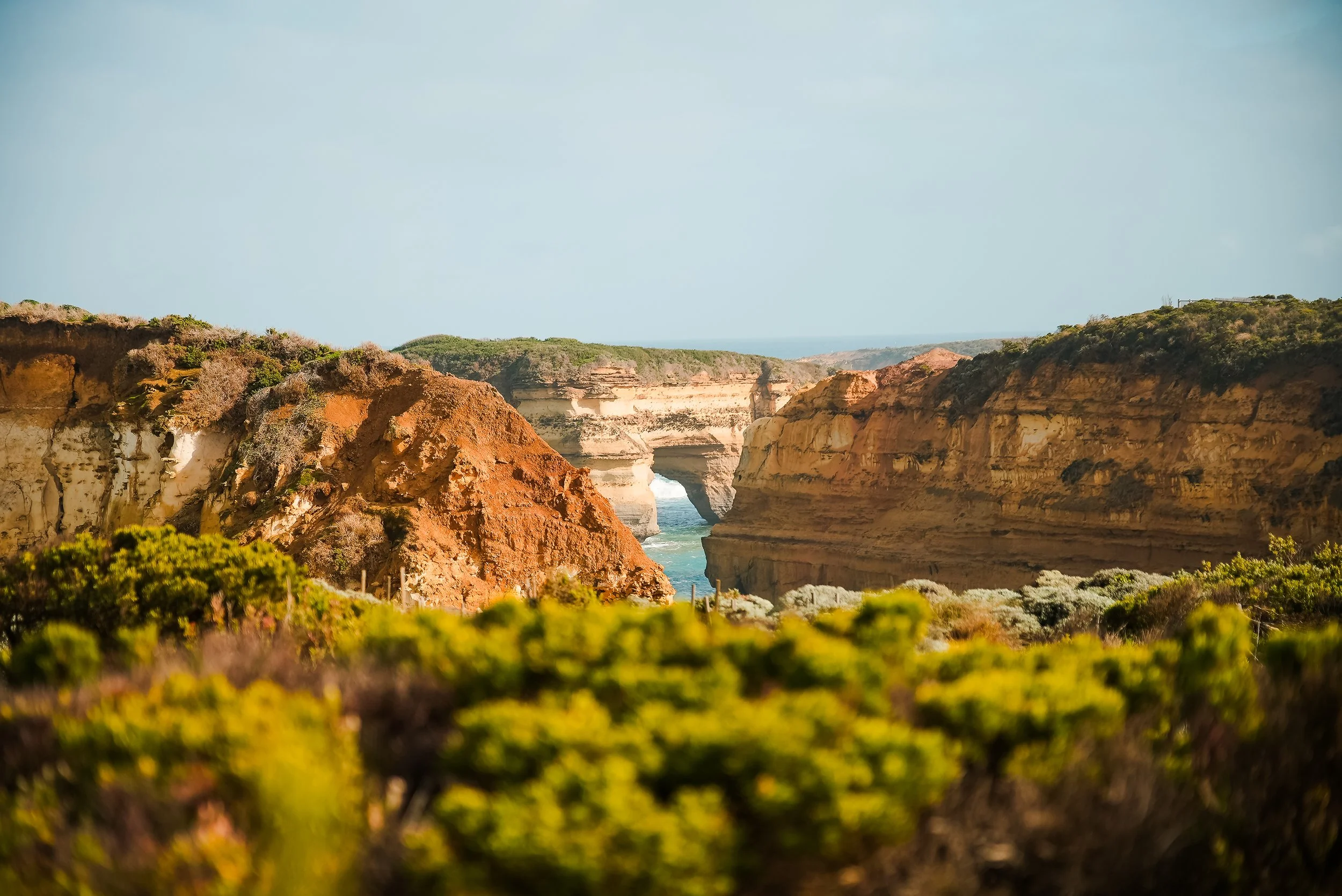 Cliffside ocean view with natural arch formation in the distance and green shrubbery in the foreground.