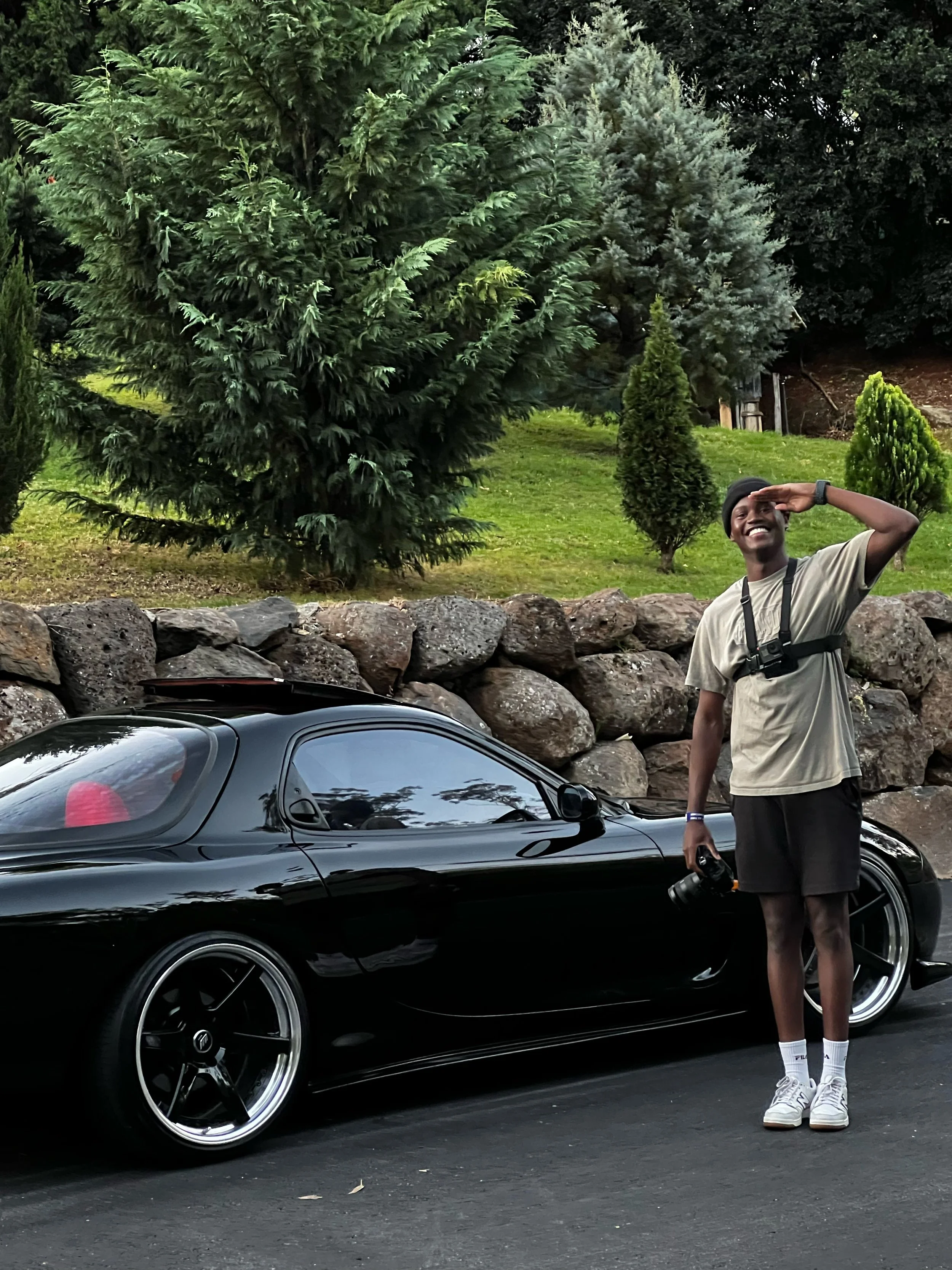 A person standing next to a black sports car on a paved road, saluting and smiling, with trees and large rocks in the background.