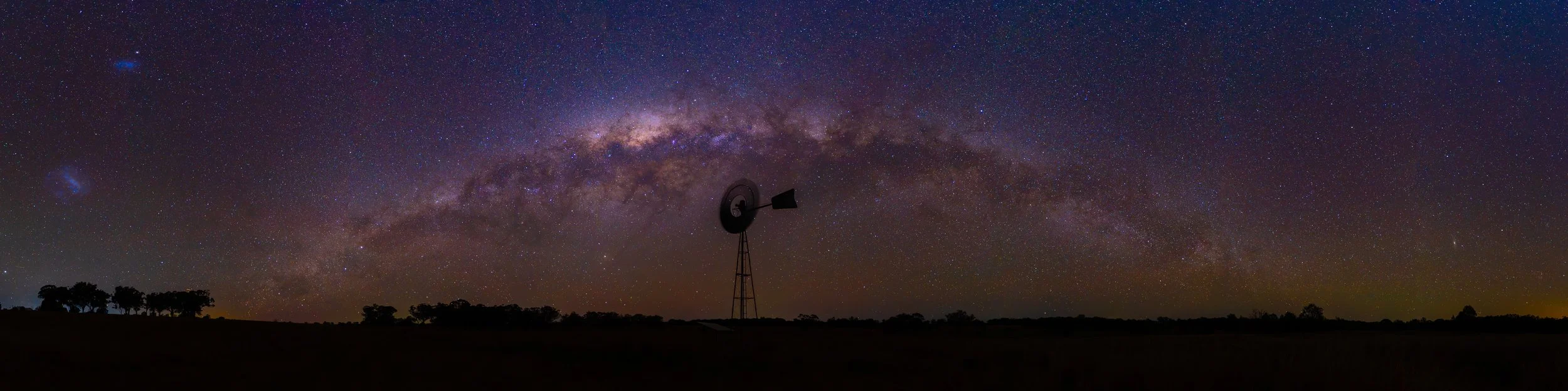 Nighttime landscape with a clear view of the Milky Way galaxy arching across the sky and a windmill silhouette in the foreground.