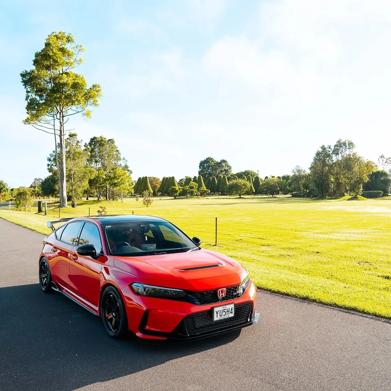 Red Honda Civic Type R parked on a paved road beside a grassy park with trees and neatly trimmed bushes under a clear blue sky.