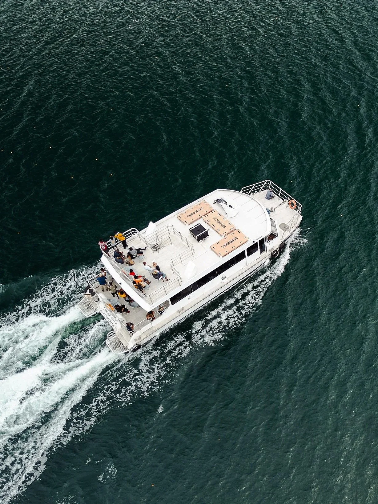 An aerial view of a white boat with multiple passengers on board, cruising through dark green water.