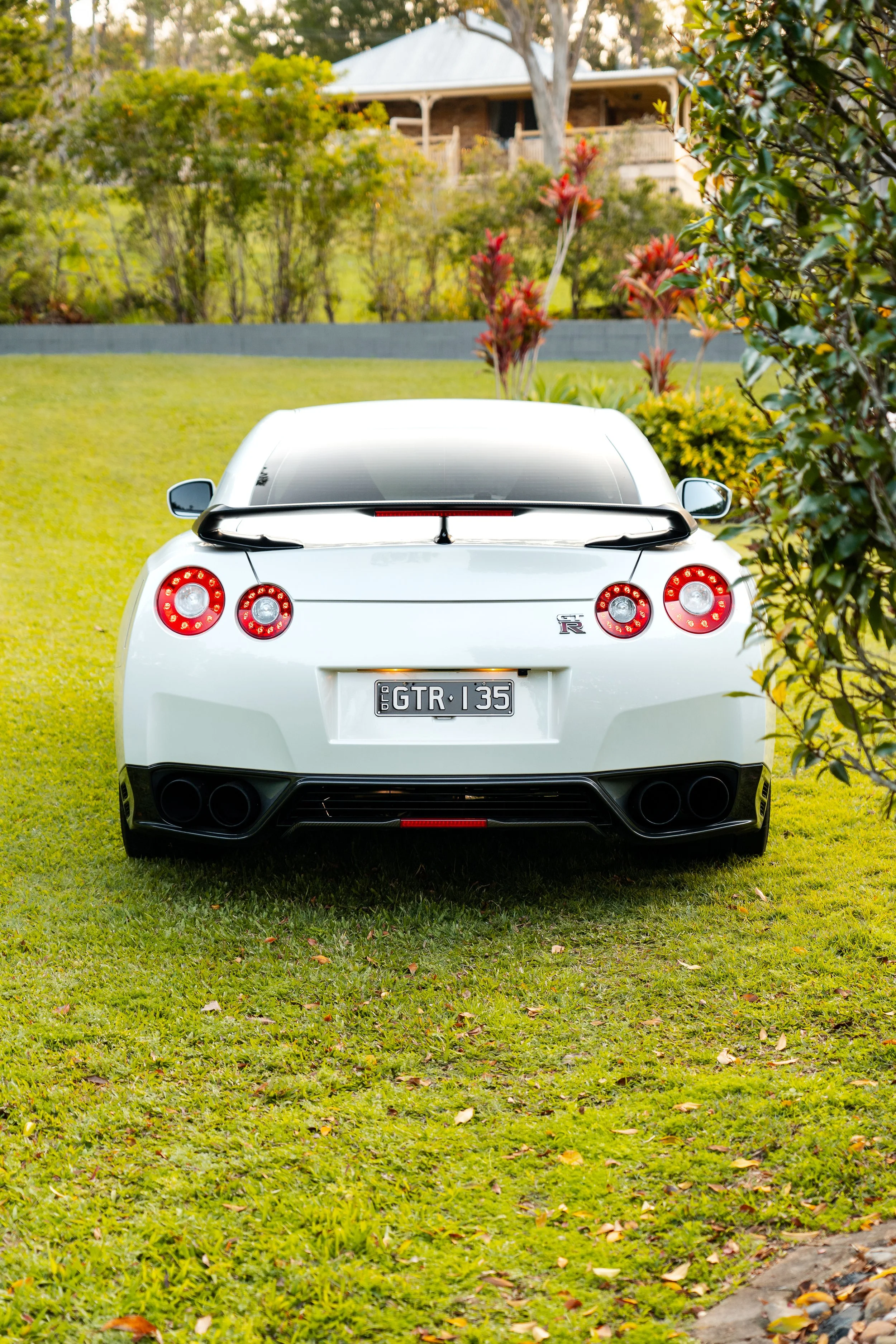Rear view of a white Nissan GT-R parked on a grassy yard, surrounded by trees and bushes, with a house visible in the background.