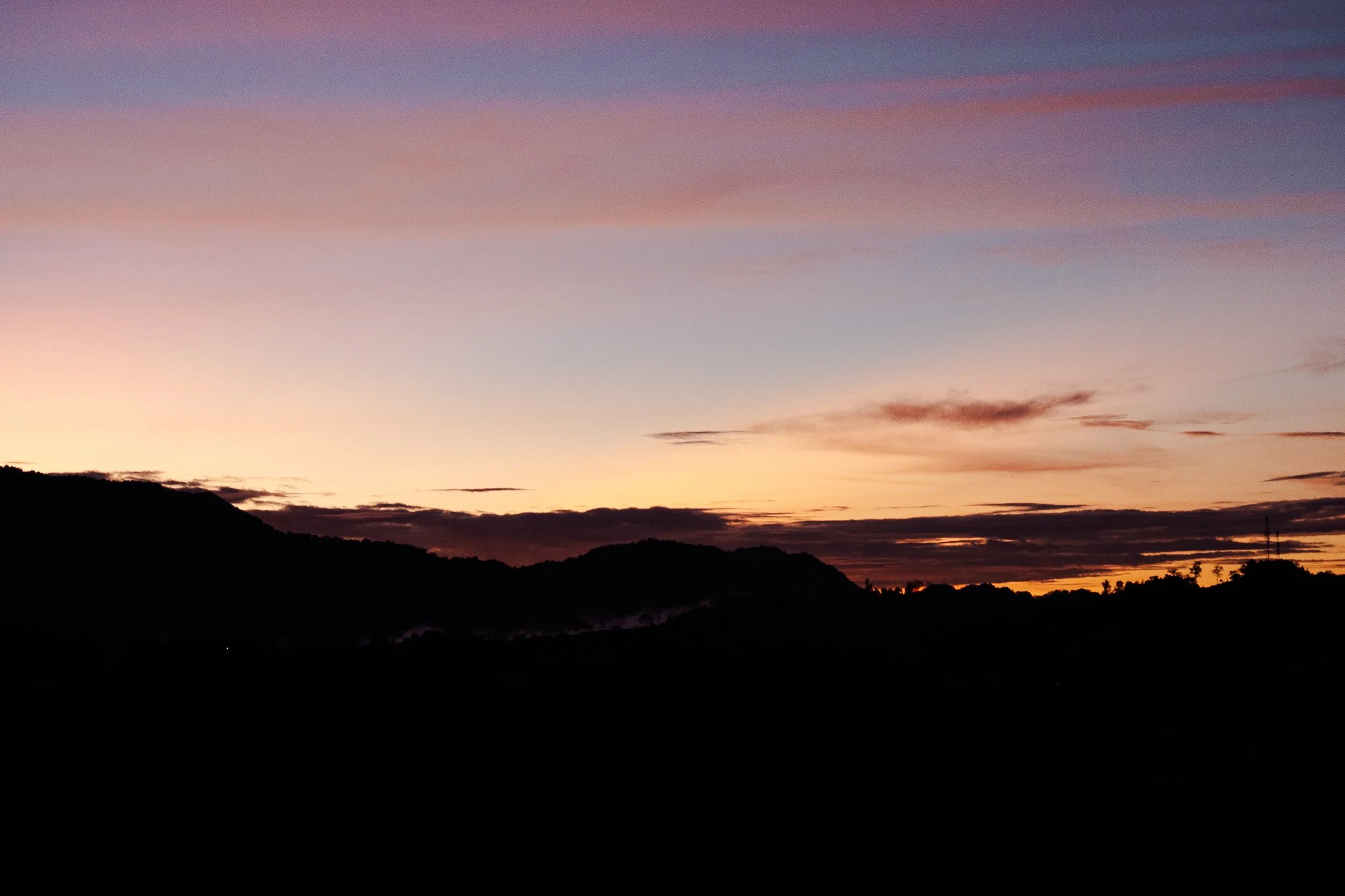 Sunset over a mountain ridge with the sky painted in shades of pink, purple, and orange, and dark silhouettes of trees and hills in the foreground.