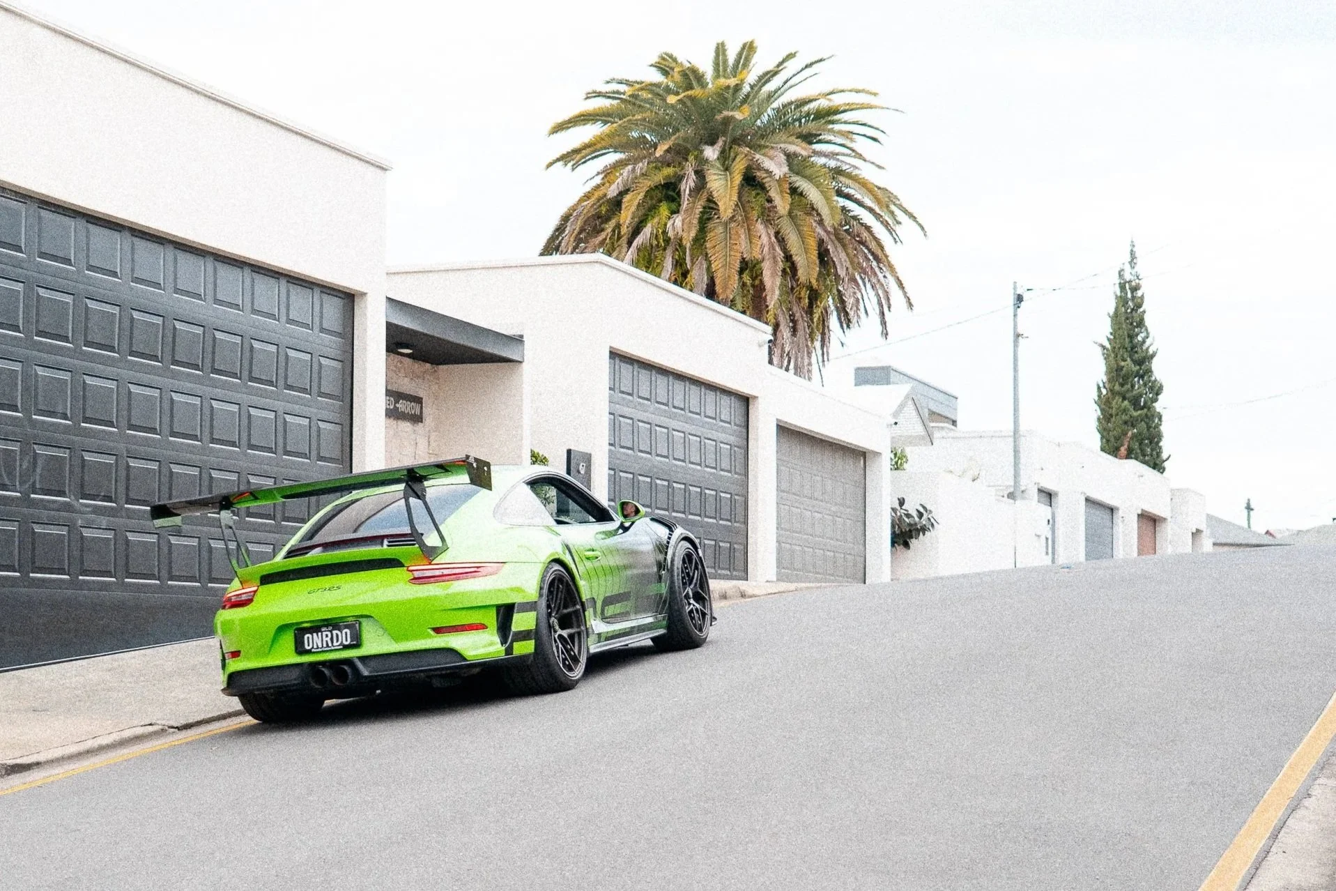 A bright green sports car with a large rear wing parked on a sloped residential street in front of modern white houses with black garage doors, featuring a tall palm tree and other trees in the background.