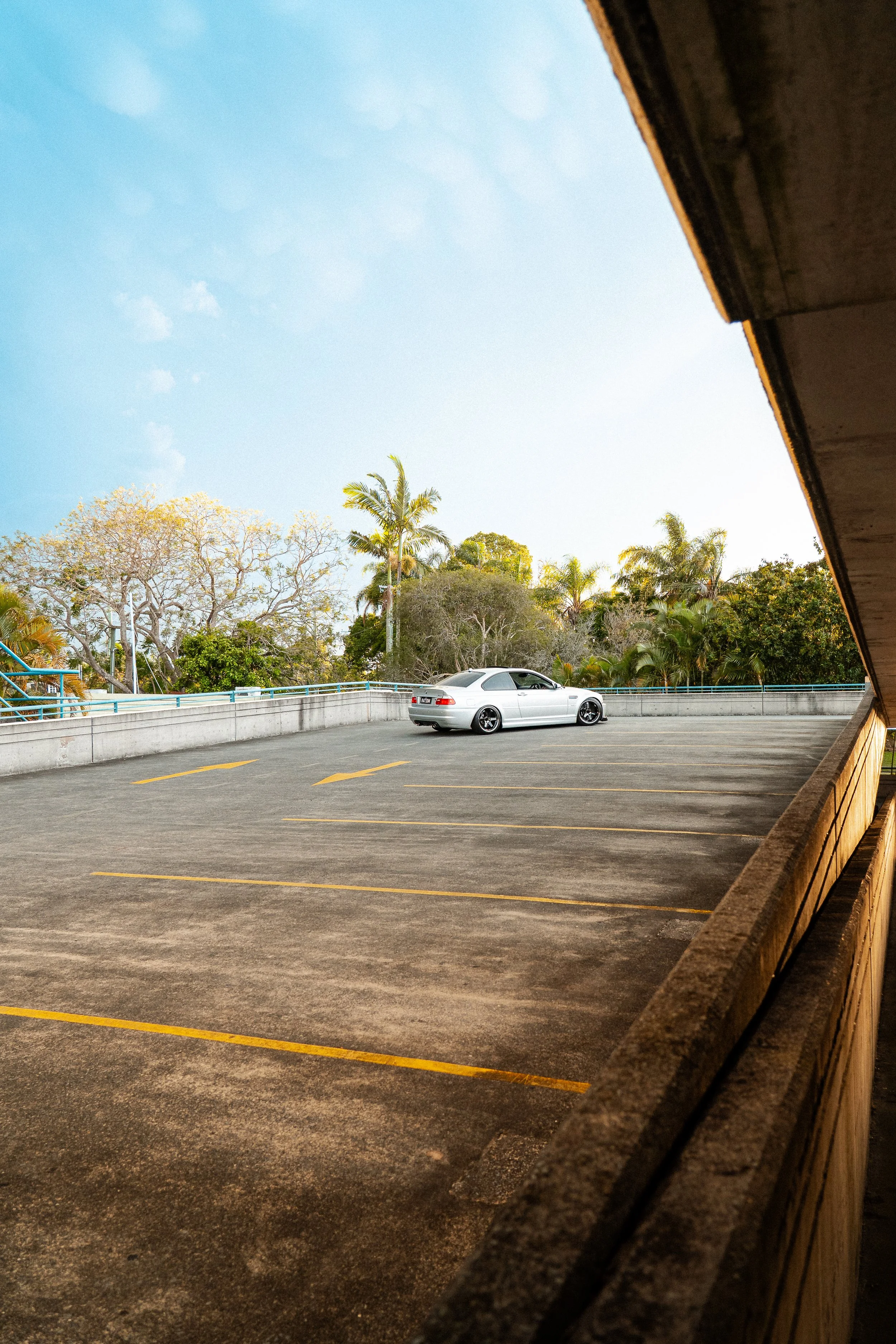 Empty parking lot with a single white car, trees with some leaves, clear sky, and a concrete railing