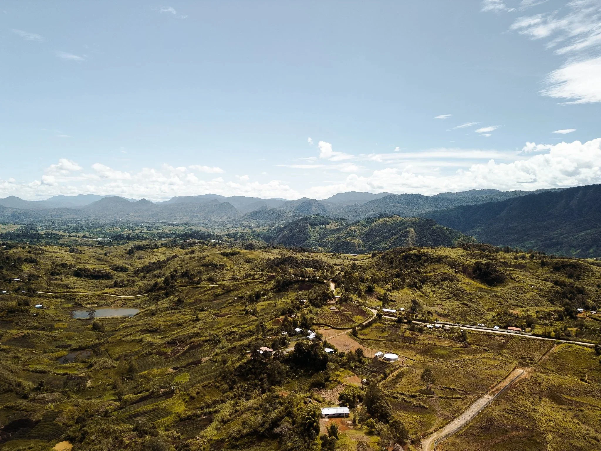 A panoramic view of a lush, mountainous landscape with rolling hills, small ponds, and scattered houses under a partly cloudy sky.