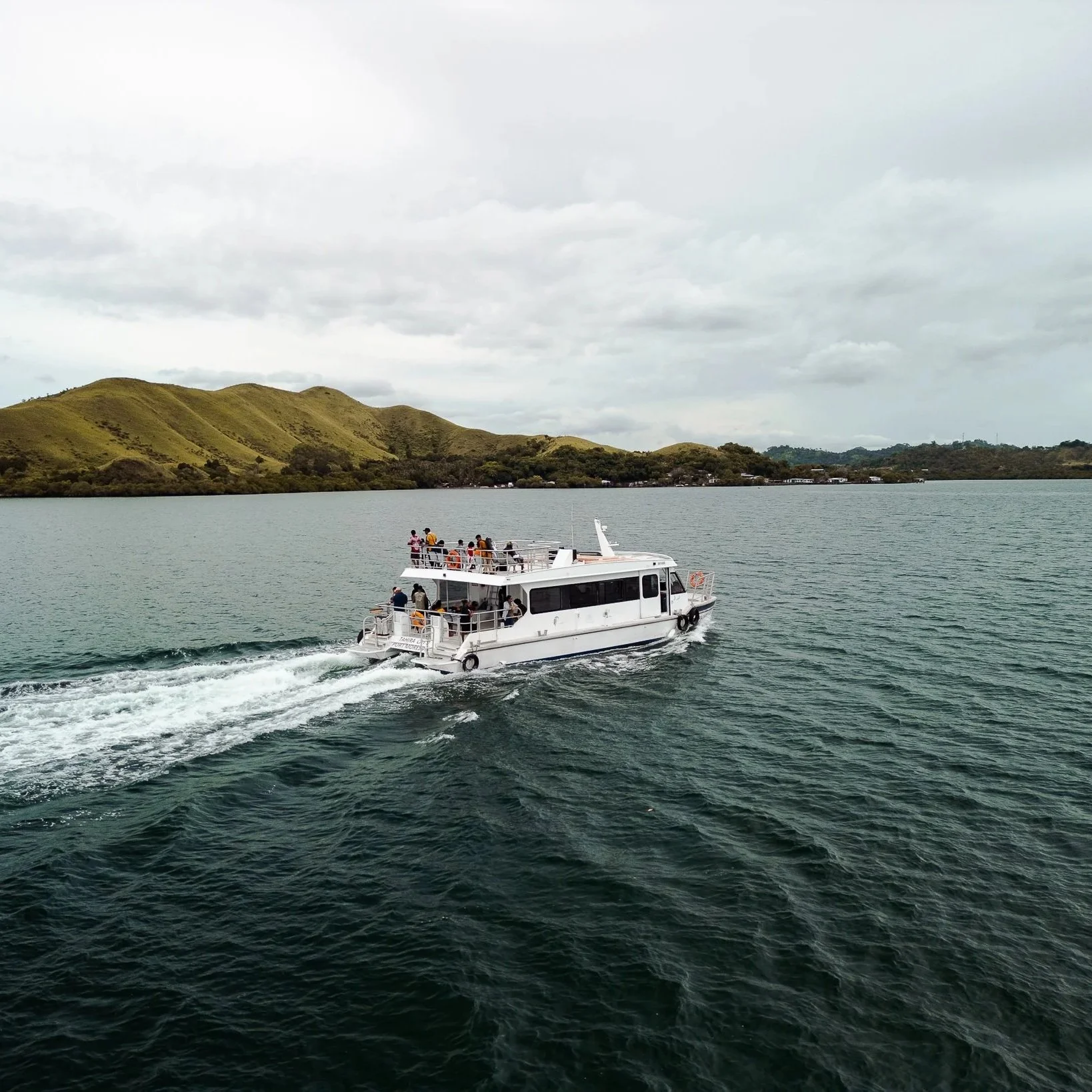 A white passenger boat cruising on calm water with a hilly landscape in the background and an overcast sky.
