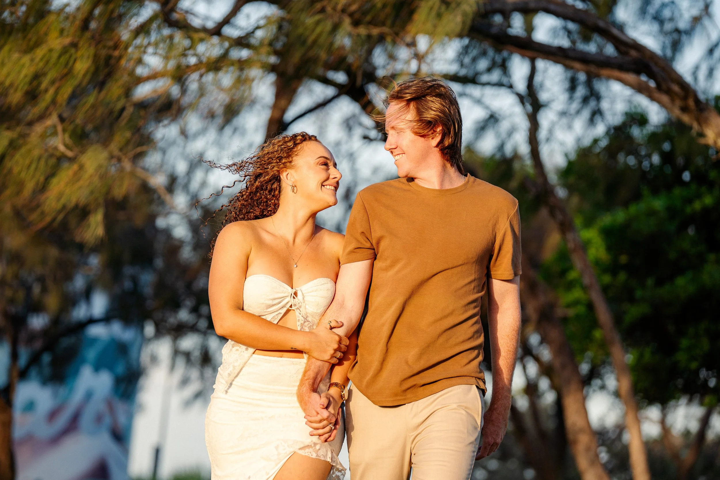 A couple walking outdoors, holding hands, smiling at each other with trees in the background.