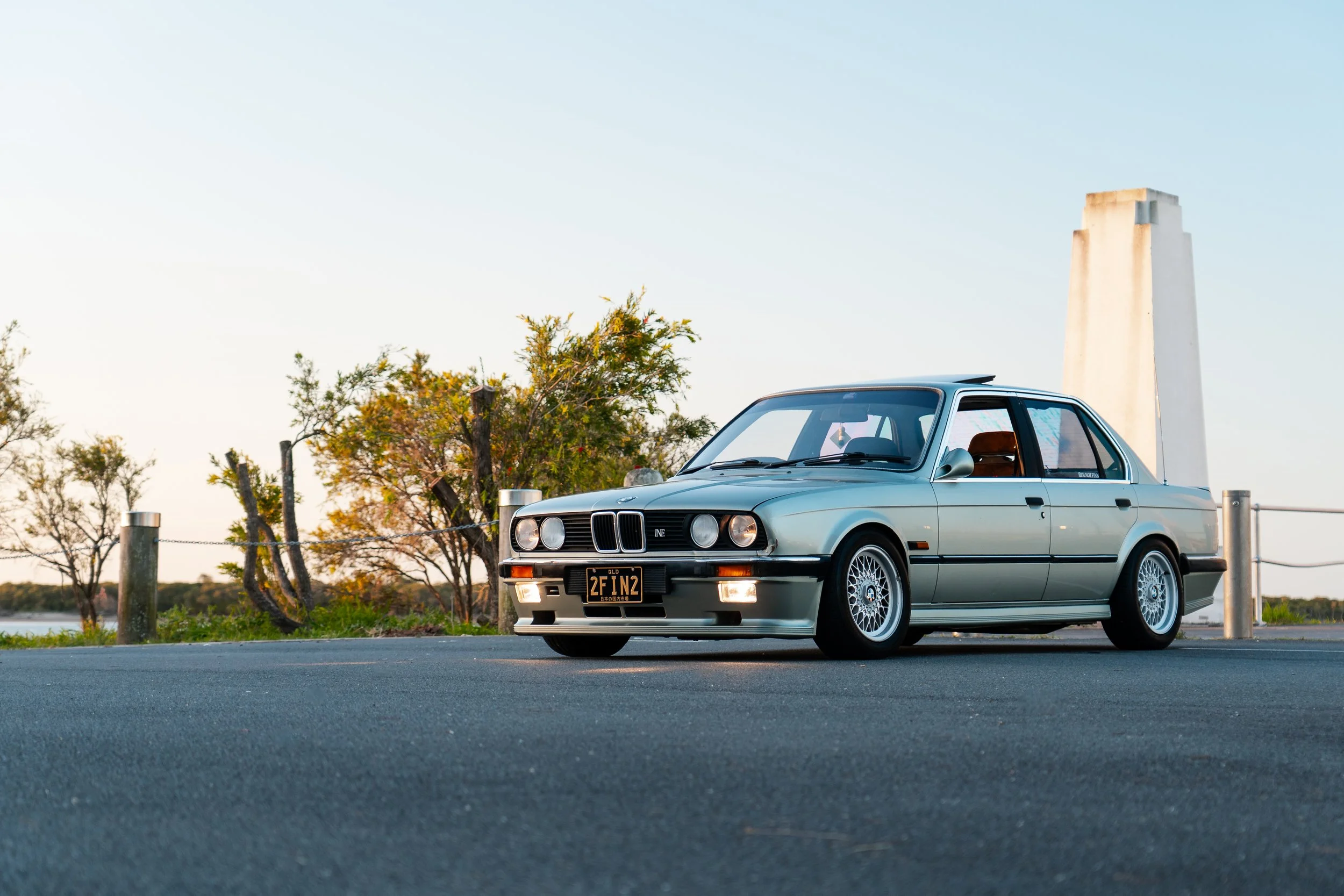 A silver classic BMW car parked on a paved surface near trees and a large white concrete structure during sunset.