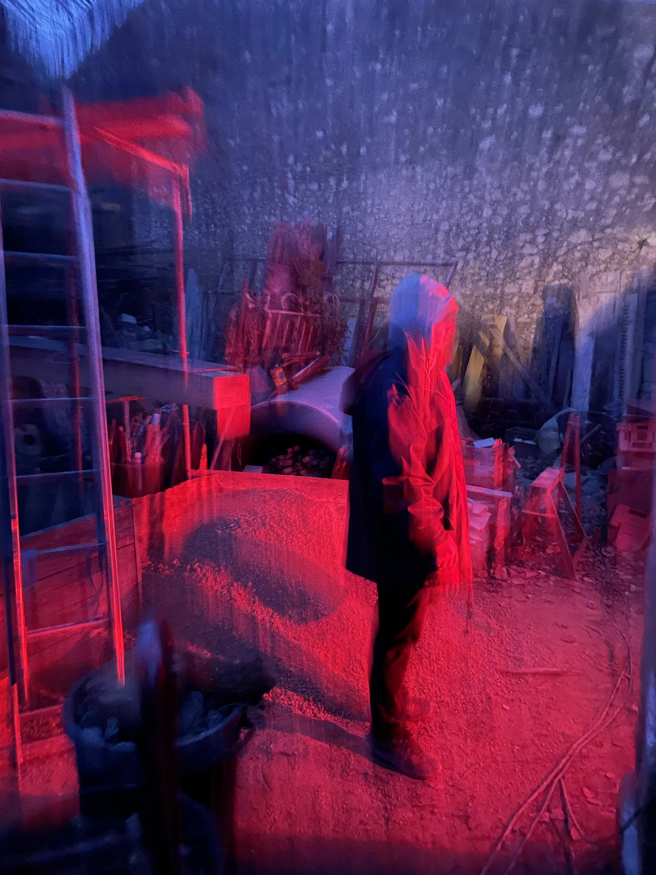 Une personne au clair de lune ou sous une lumière rouge, debout dans un atelier ou une zone de stockage, entourée de meubles, outils et matériaux divers.