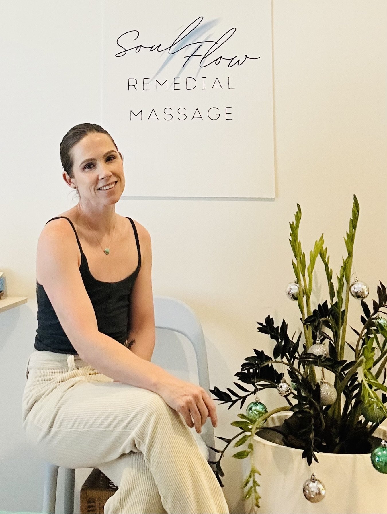 A woman sitting on a chair at a remidal massage center with a sign that reads 'Soul Flow Remedial Massage' behind her and holiday ornaments hanging on a potted plant.