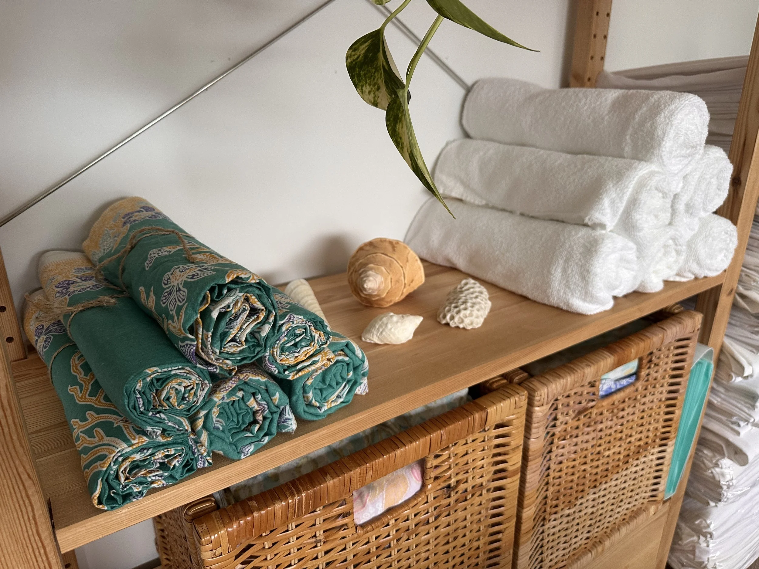 A wooden shelf contains neatly rolled towels, seashells, and a plant leaf. The top shelf has six rolled white towels stacked and four seashells. The bottom shelf has three folded patterned fabric pieces.