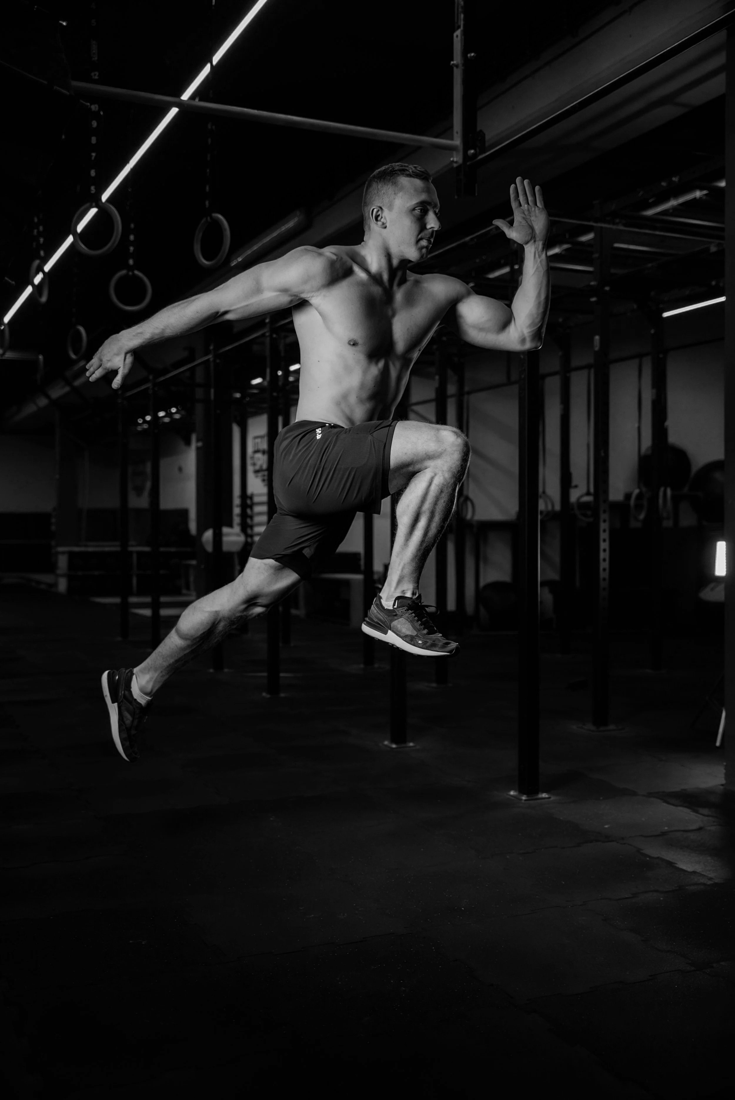 Ein fitter Mann trainiert im Fitnessstudio, springt in die Luft, hebt die Arme und ist nur in Performance Sportshorts und Turnschuhen gekleidet. Das Bild ist in Schwarz-Weiß. RUSH IT. Sporthose