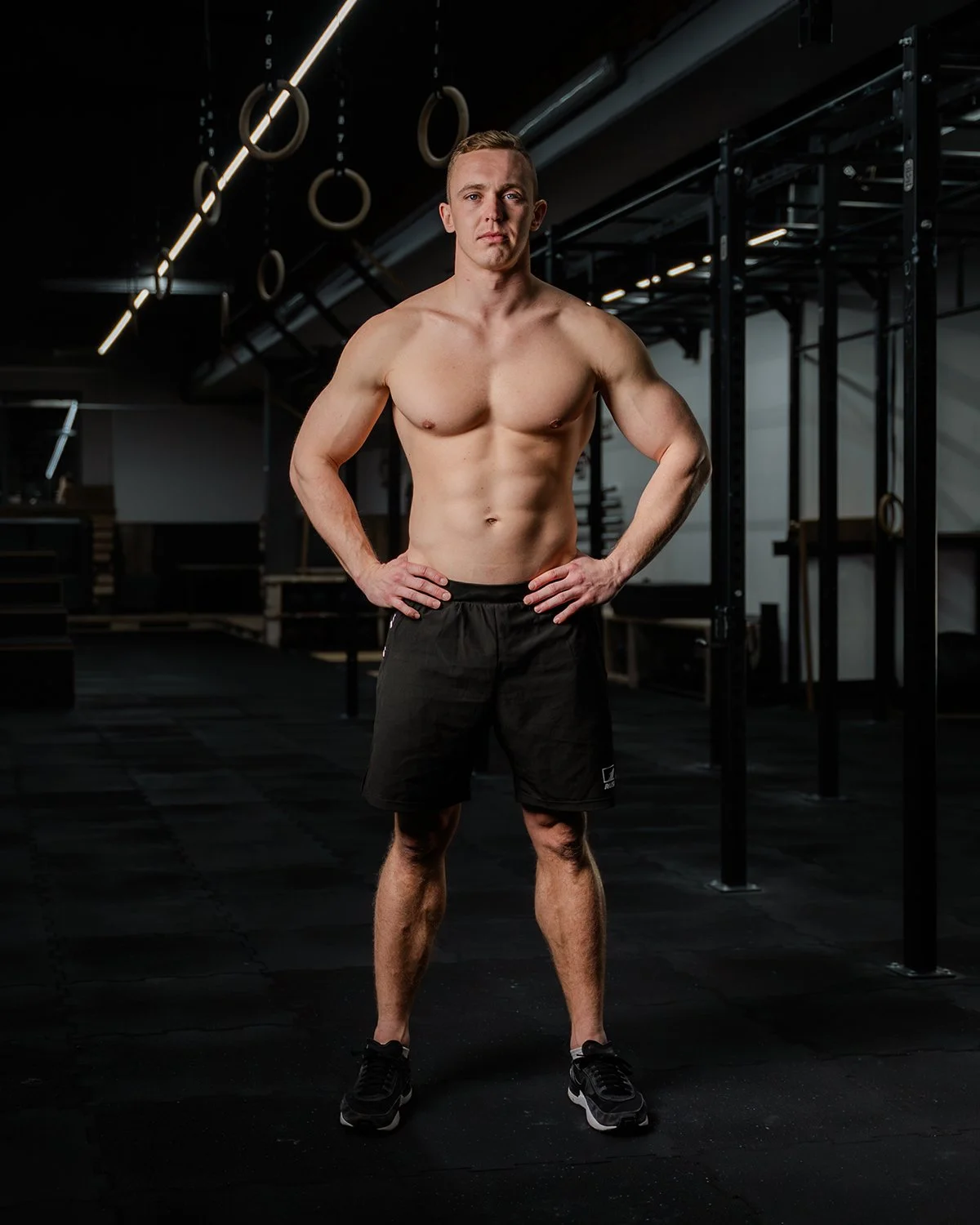 Ein junger, muskulöser Mann steht sportlich in einem Fitnessstudio mit schwarzen Turnschuhen, schwarzer Performance Sportshorts und Position mit den Händen auf den Hüften, vor schwarzem Trainingsgerüst und Turnringen im dunklen Raum.  RUSH IT.