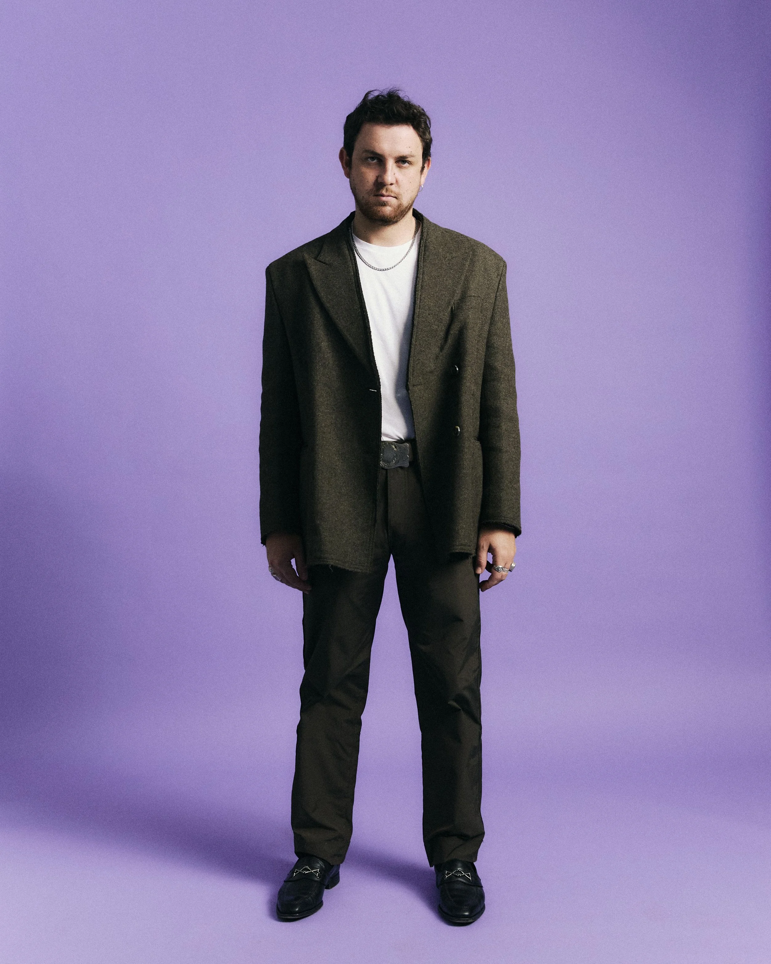 A man standing against a purple background, wearing a dark blazer, white t-shirt, black pants, black shoes, and jewelry.