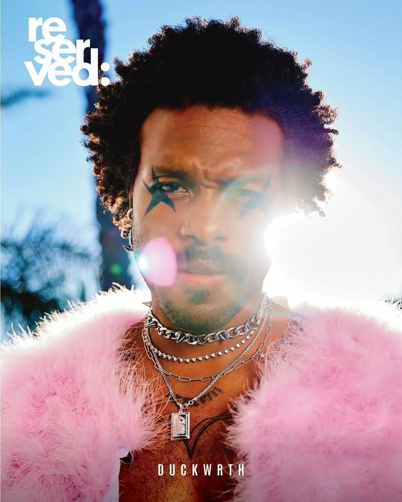 Close-up photograph of a person with dark curly hair, wearing multiple silver necklaces, earrings, and a pink fluffy garment, with face makeup resembling animal features, standing outdoors in sunlight with a lens flare.