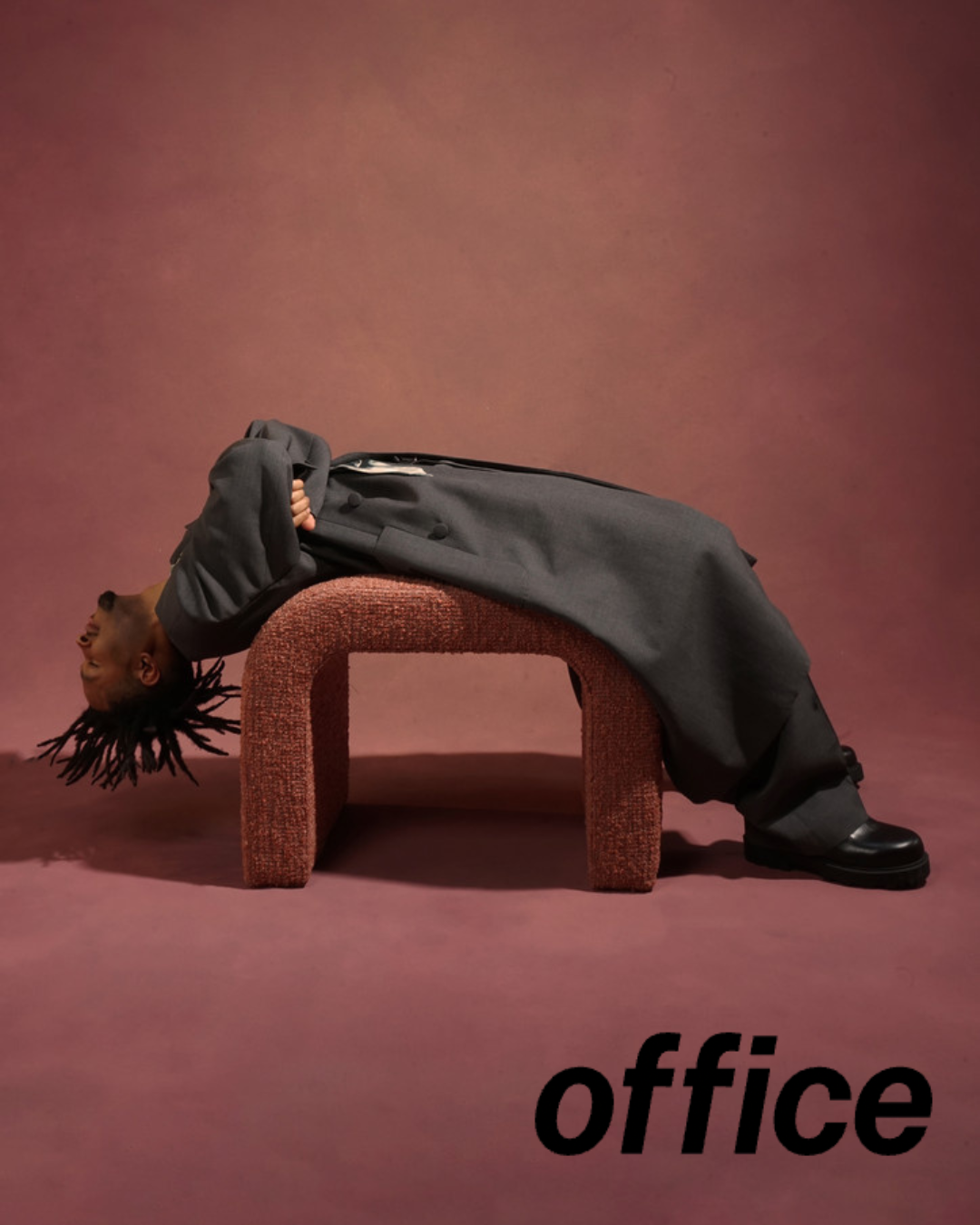 A person with dreadlocks lying back on a small red upholstered stool, with head hanging upside down and smiling, wearing a gray coat and black shoes, against a pink background. The word 'office' is written in black at the bottom right corner.