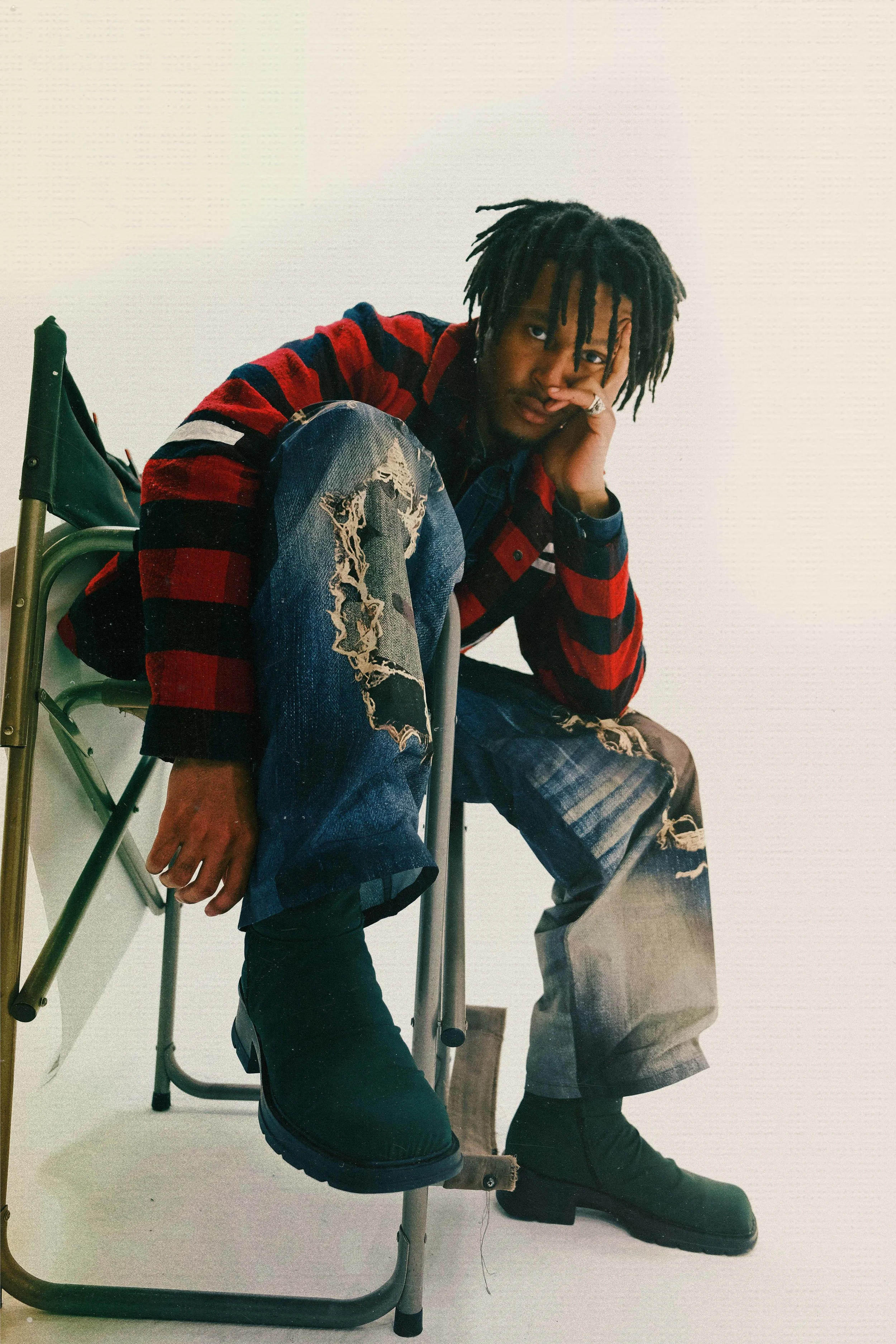 A young man with dreadlocks sitting on a chair, resting his head on his hand, wearing a red and black striped jacket, ripped jeans, and black boots, against a plain white background.